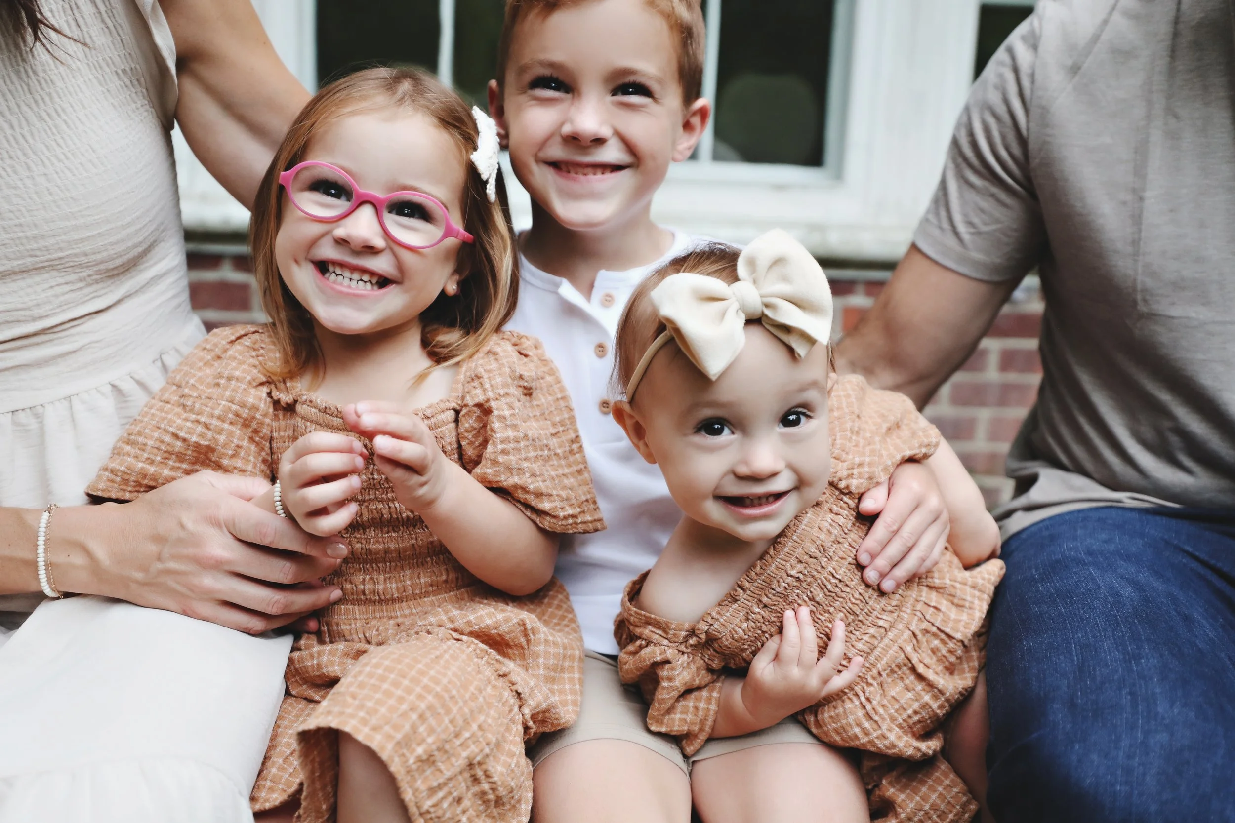 A family with two young girls sitting outdoors, smiling and hugging each other. The girls are wearing matching brown dresses, and one has pink glasses and the other has a beige bow headband. A boy in a white shirt is sitting between them, smiling.