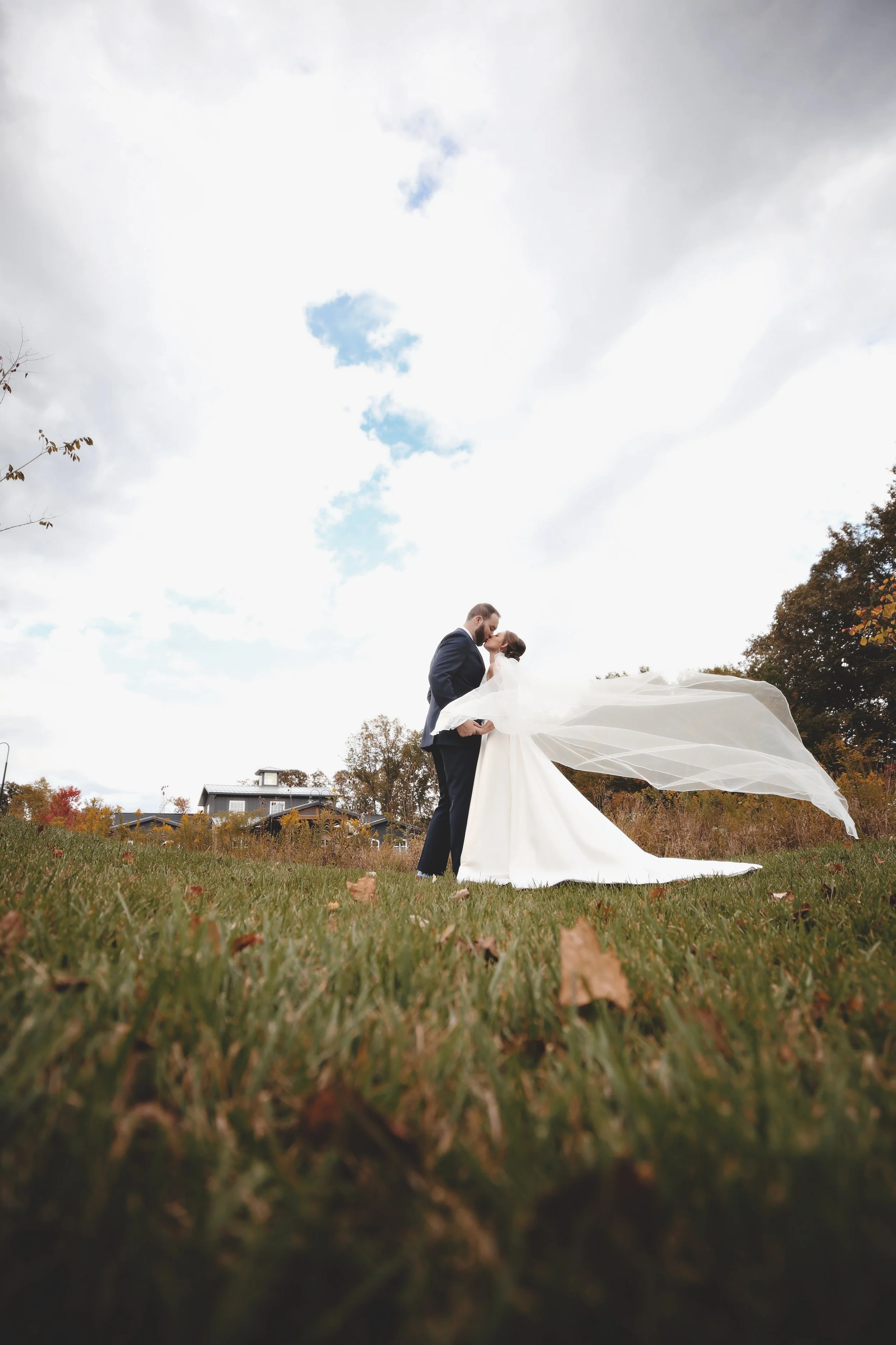 A newlywed couple sharing a kiss outdoors, with the bride in a long white gown and veil, and the groom in a dark suit, standing on grass with autumn leaves, trees, and a house in the background under a partly cloudy sky.