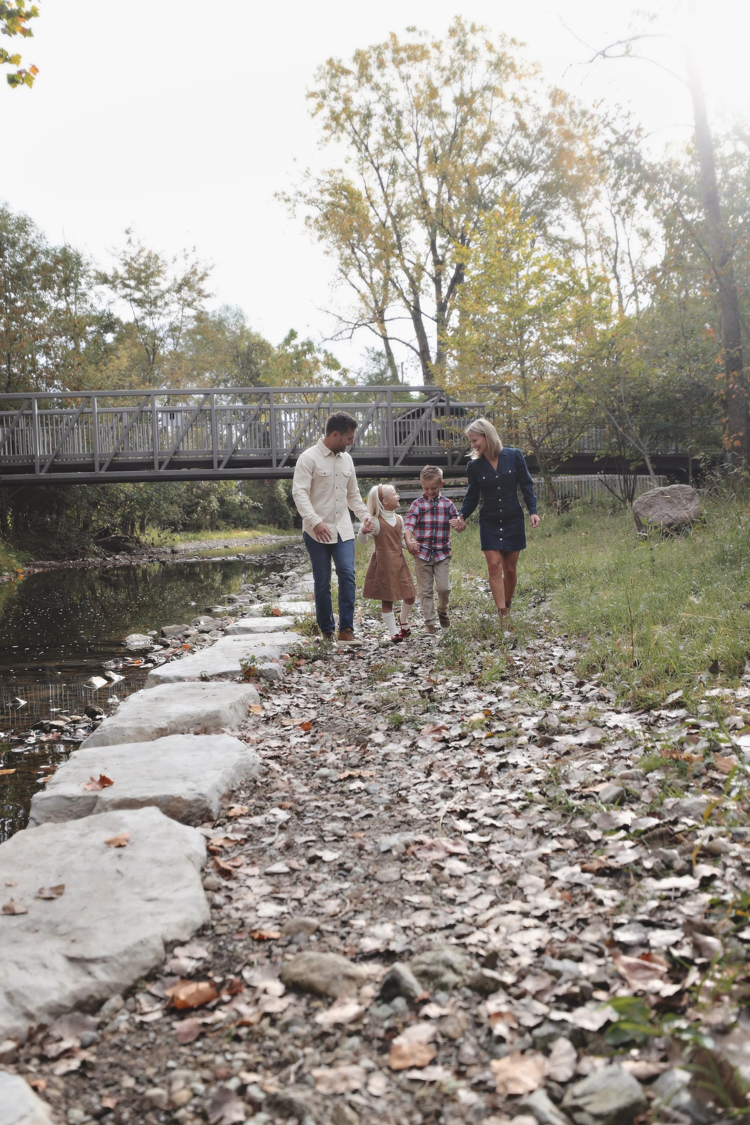 A family of four walking on a rocky, leaf-covered trail by a creek, with trees and a footbridge in the background on a clear day.