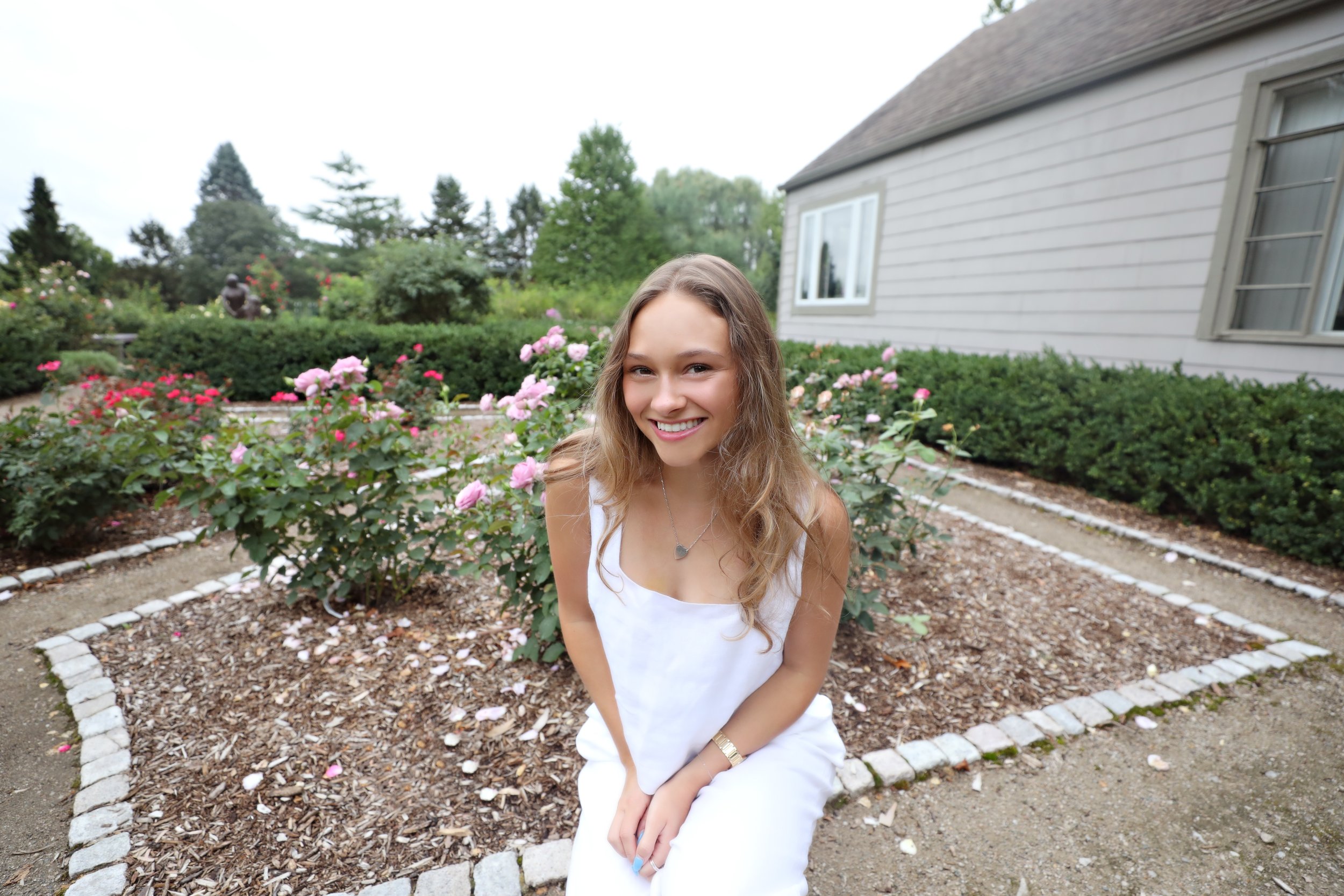A young woman with long wavy hair smiling and sitting in a garden with pink and white roses, in front of a house with gray siding and windows.