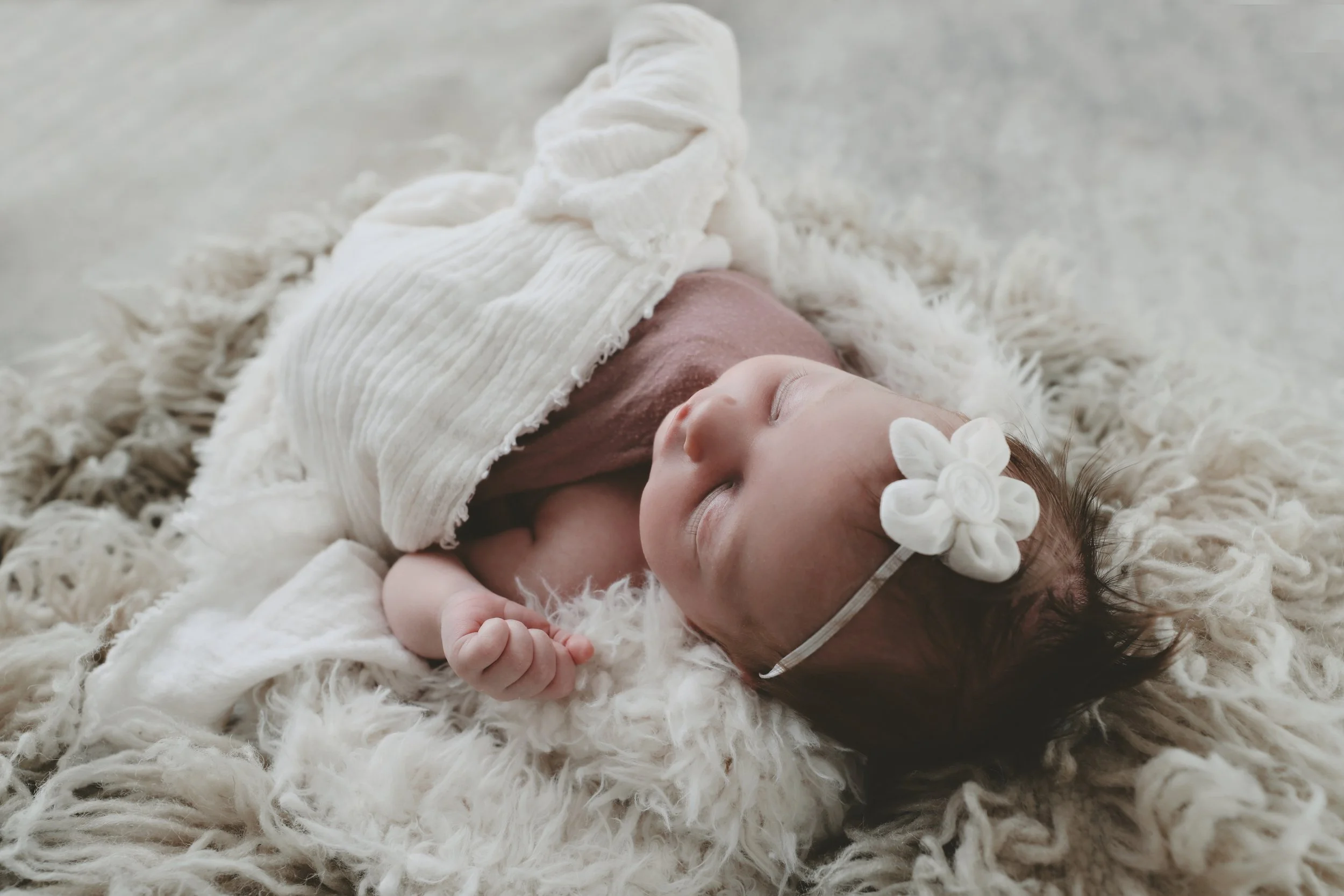 A sleeping baby girl lying on a fluffy white blanket, wearing a white headband with a fabric flower and swaddled in a soft blanket.