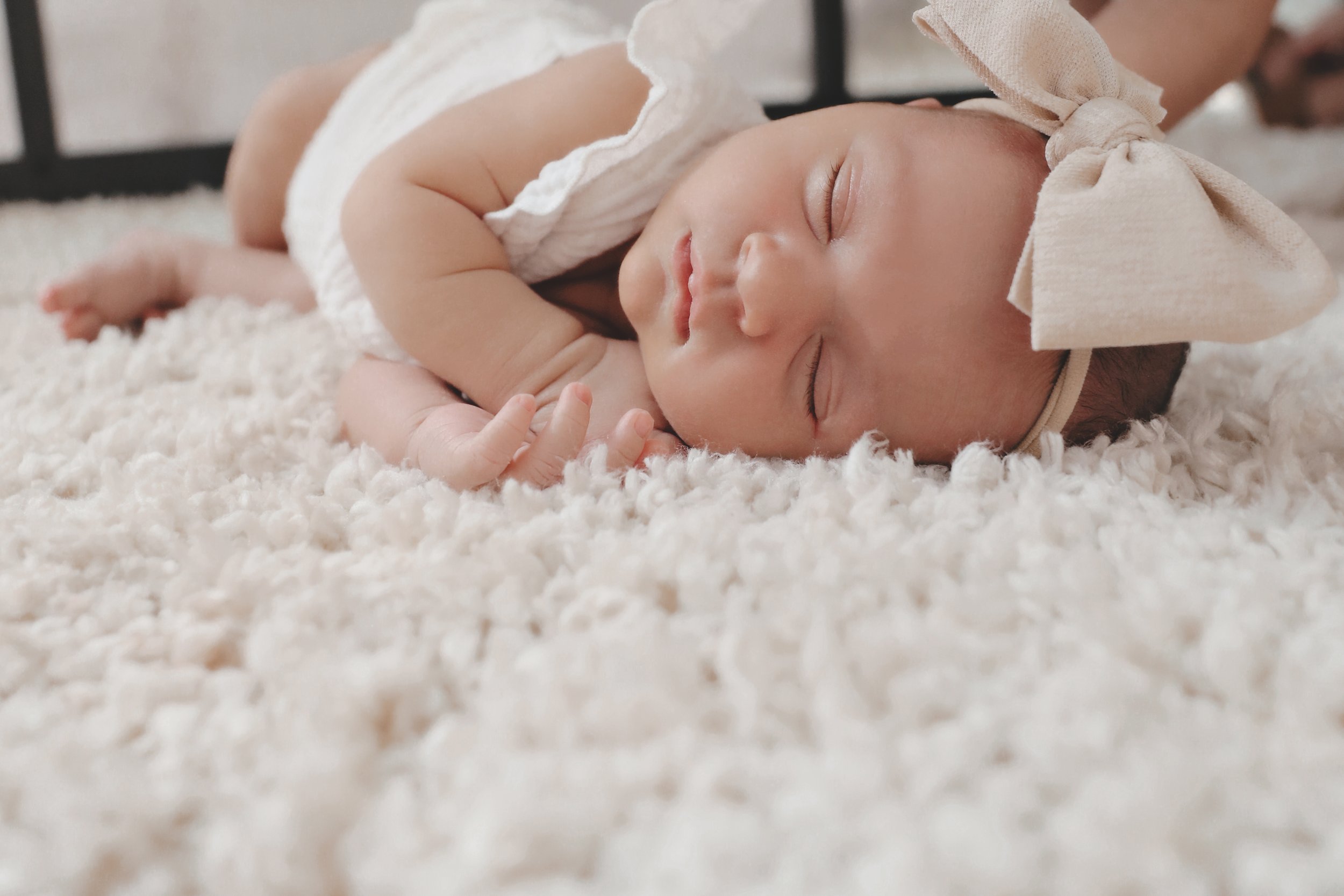 A newborn baby girl sleeping on a soft white fluffy rug, wearing a beige headband with a bow and a white outfit.