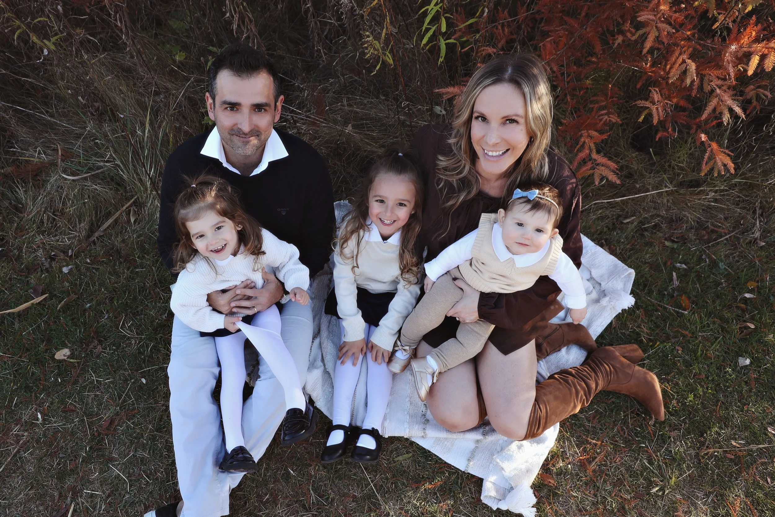 A family of five sitting on a blanket outdoors, smiling at the camera. The group includes a man, a woman, and three young girls, surrounded by autumn foliage.