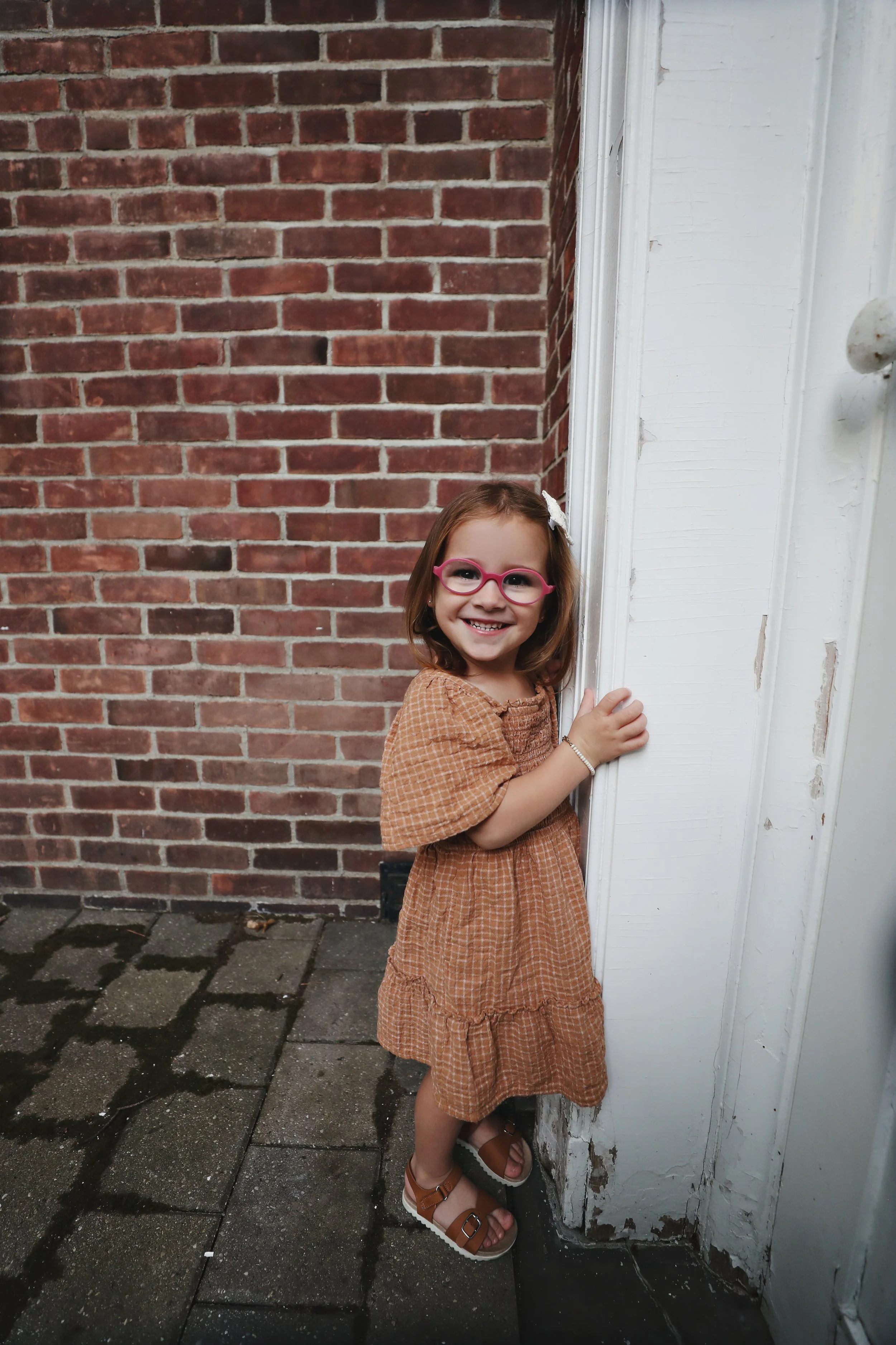 A young girl with brown hair, pink glasses, and a white bow wearing a brown checkered dress and sandals, smiling and standing next to a white doorframe against a brick wall.
