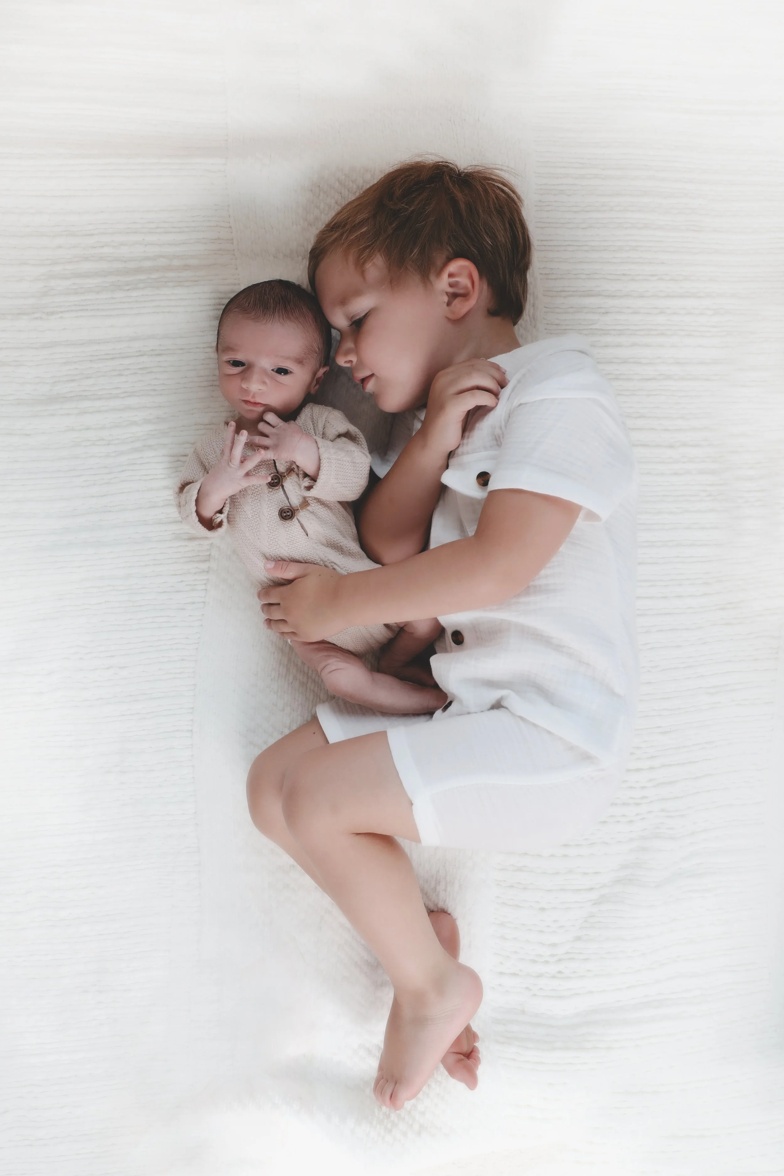 A young boy cuddling a newborn baby on a white bed, both dressed in light-colored clothing.