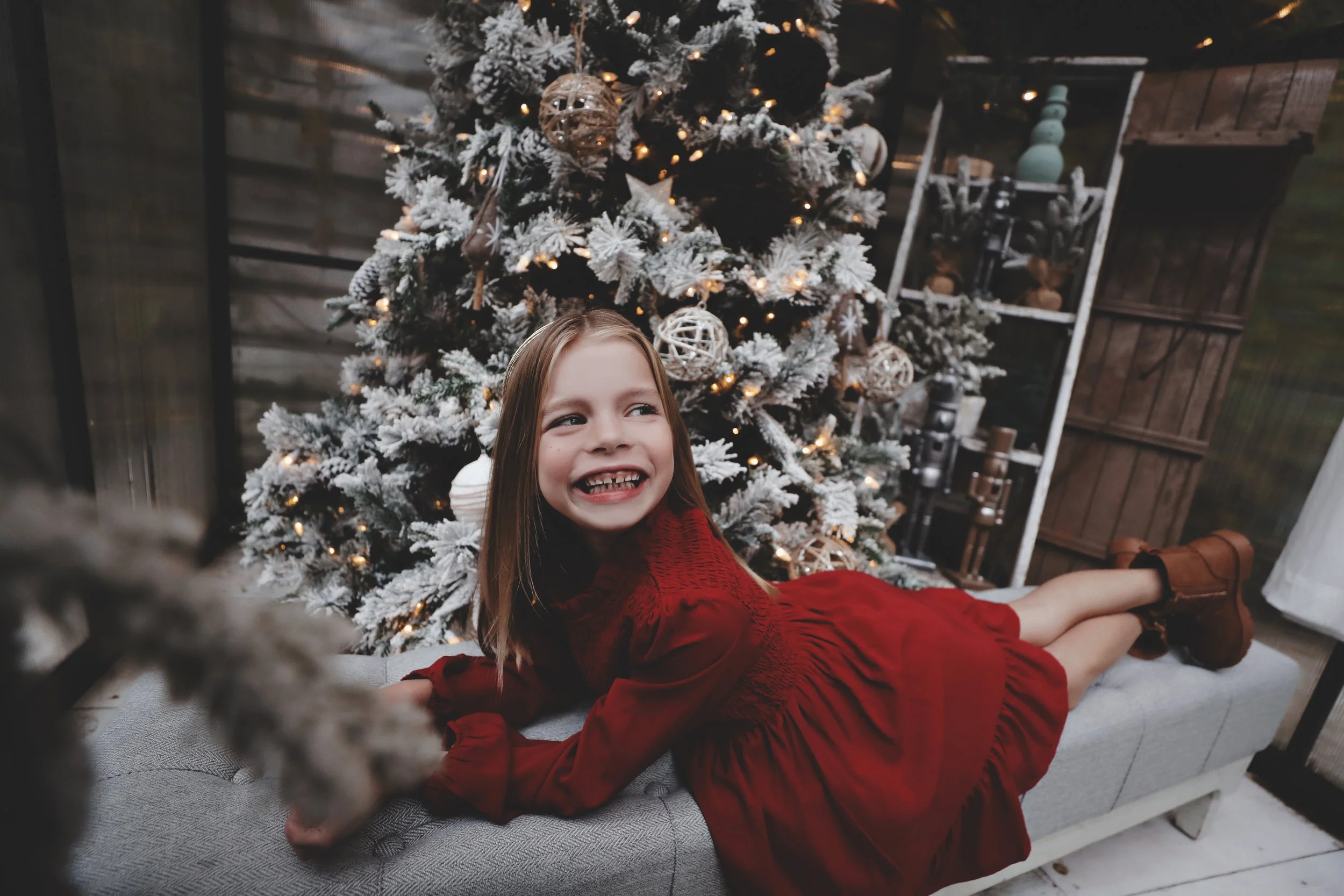 A young girl in a red dress lying on a cushioned bench in front of a decorated Christmas tree, smiling at the camera.