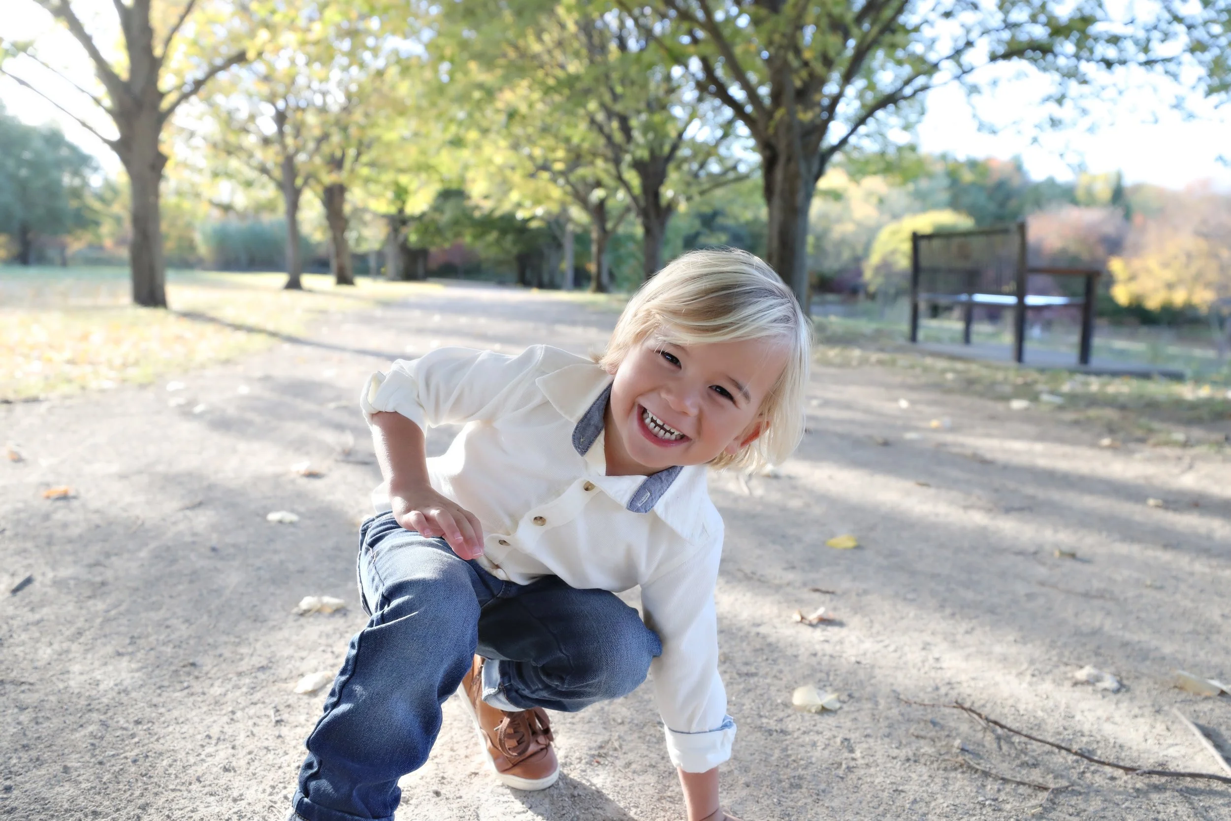 Young boy crouching and smiling outdoors in a park with trees and sunlight