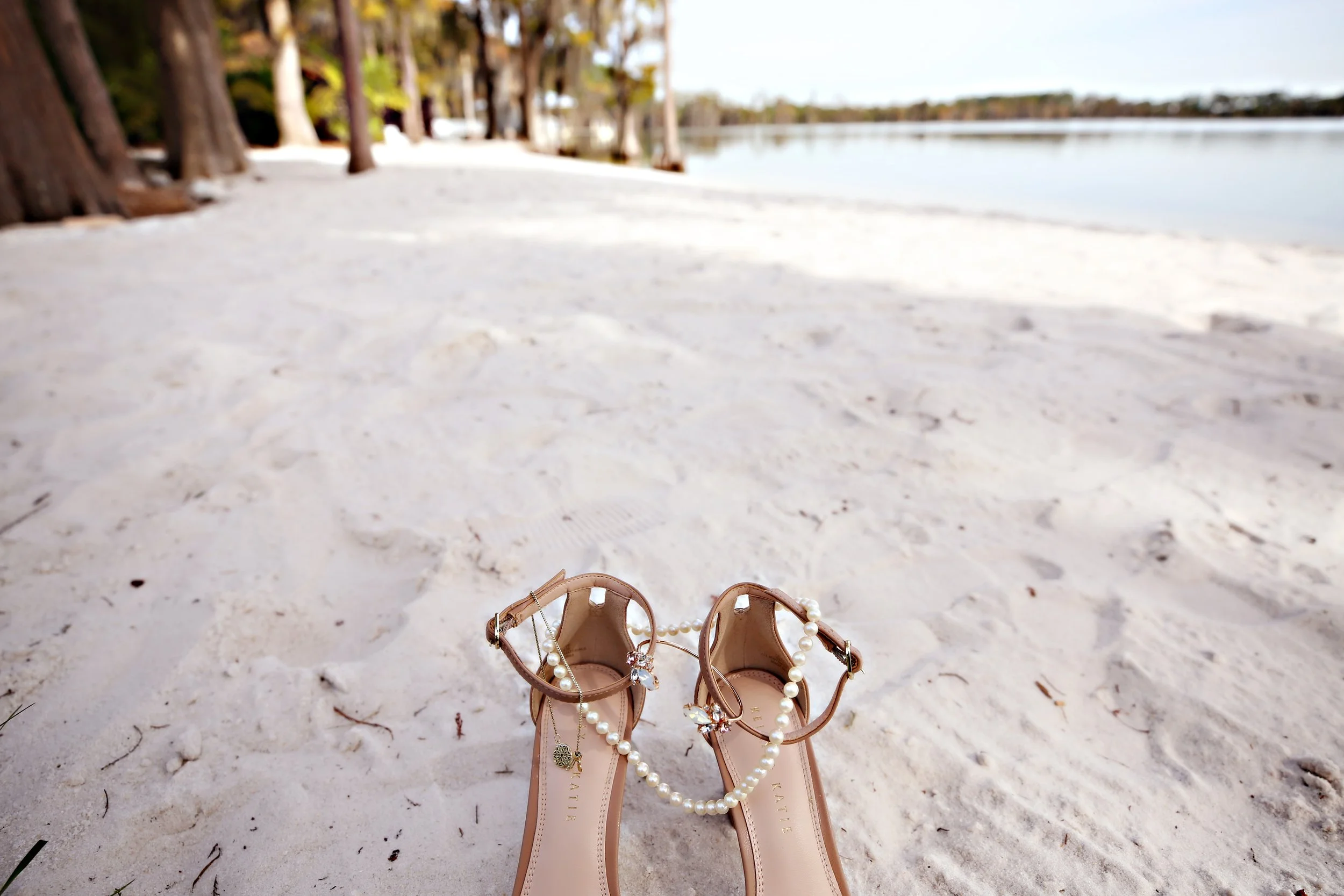 A pair of nude sandals with ankle straps, decorated with pearl and jewel embellishments, sitting on sandy beach with trees and water in the background.