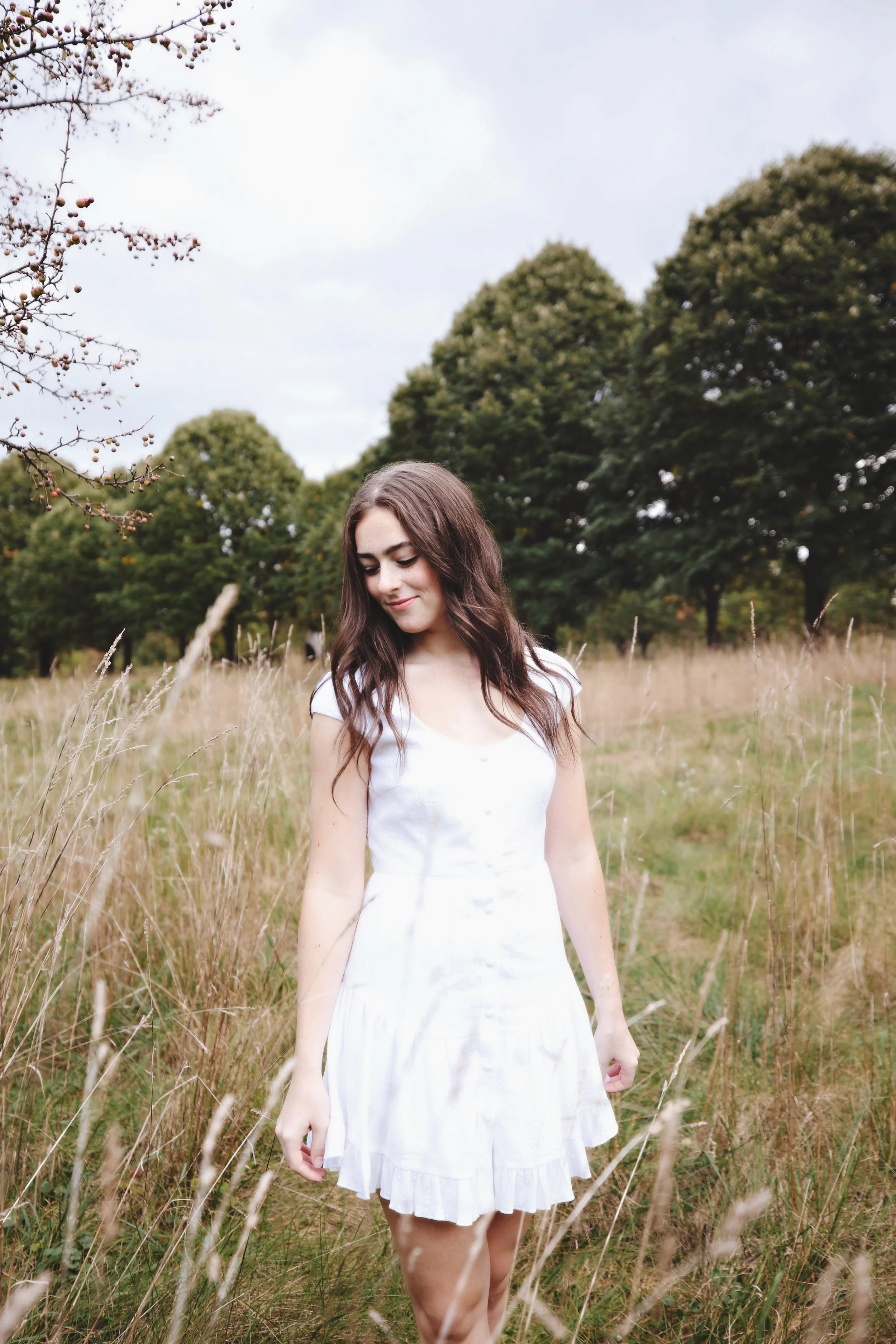 Young woman with long brown hair wearing a white dress standing in a grassy field with trees in the background.
