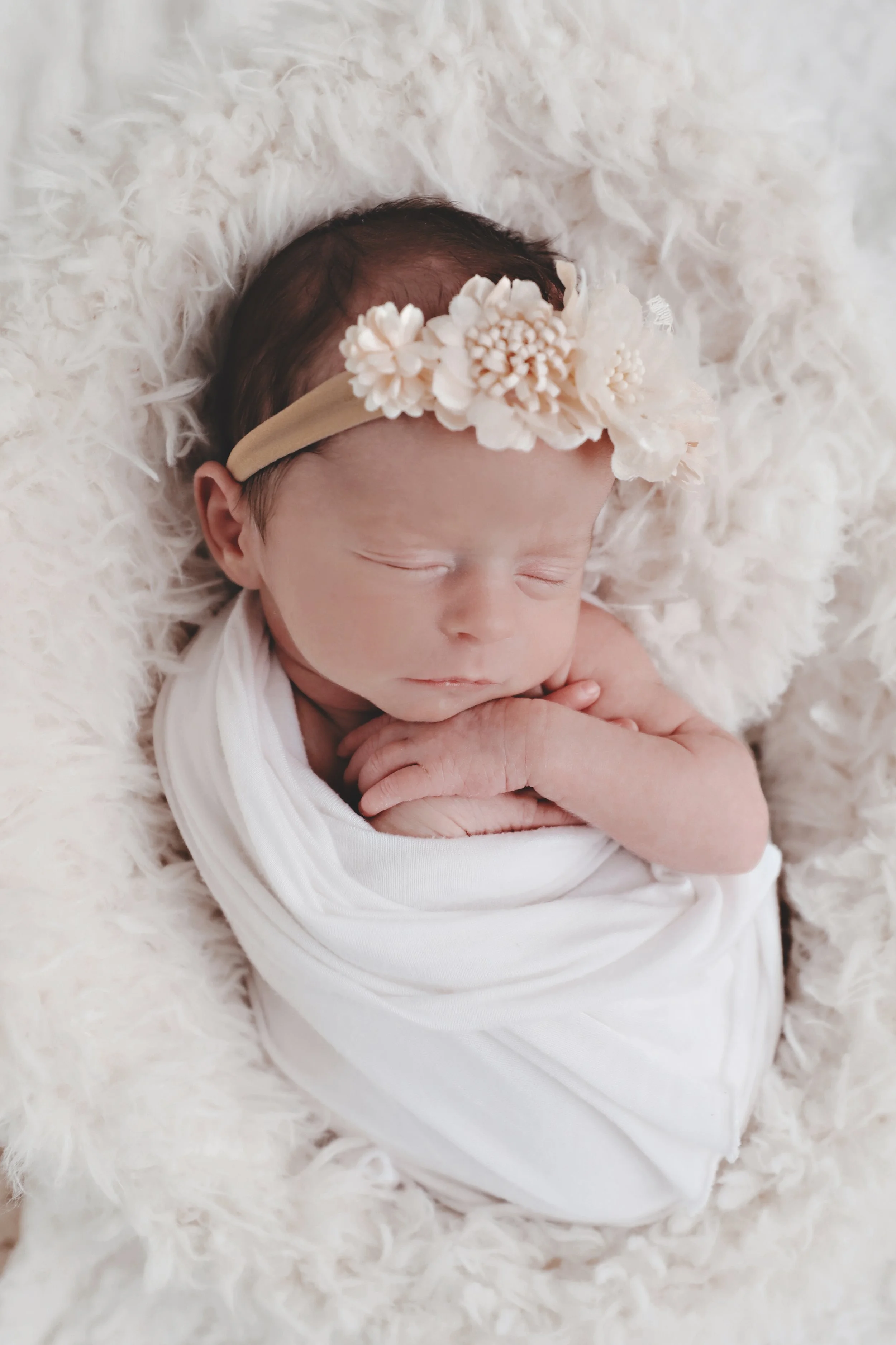 Close-up of a sleeping newborn baby wrapped in a white blanket, wearing a cream floral headband, lying on a fluffy white surface.