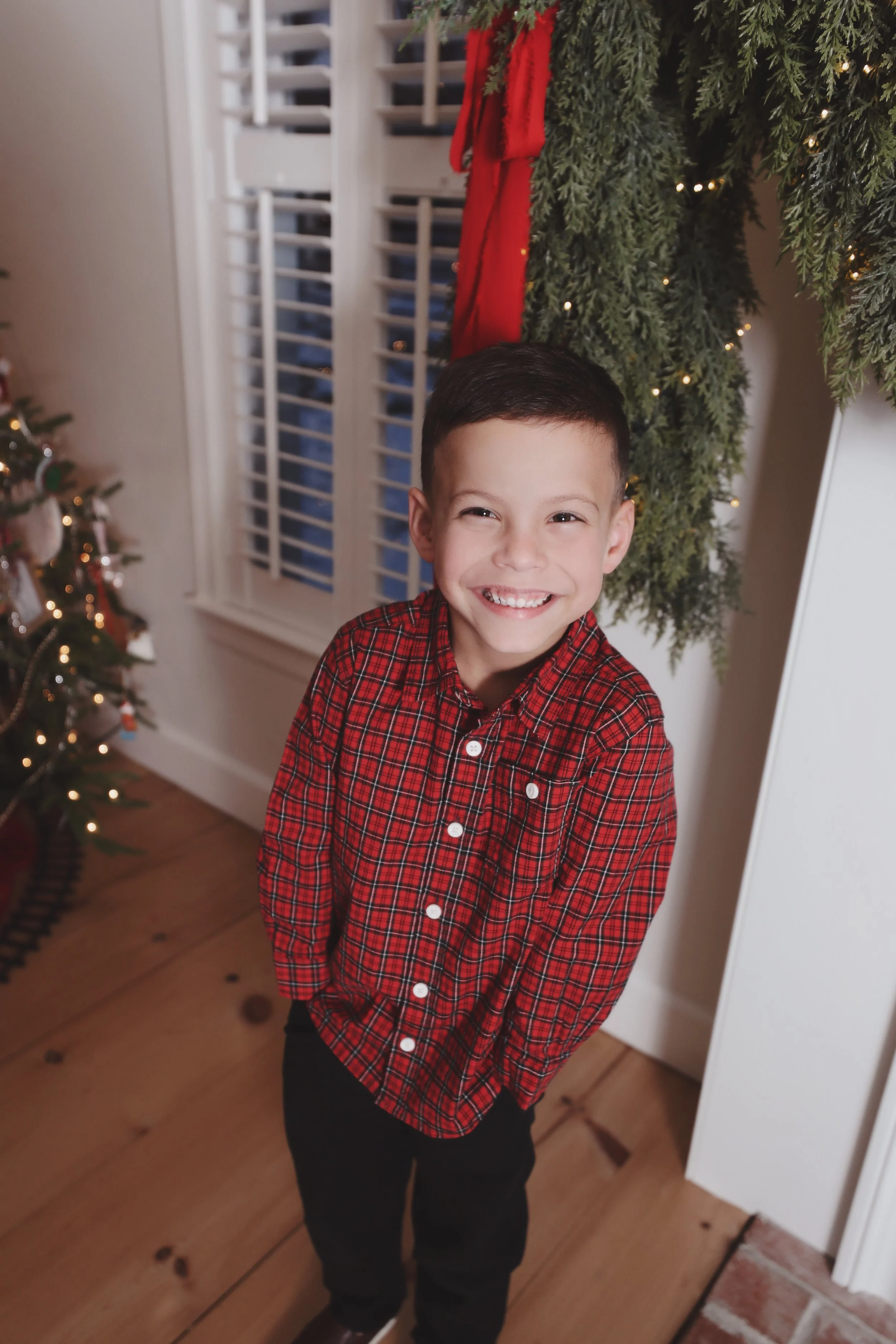 A young boy with a big smile, wearing a red plaid shirt, standing indoors near a Christmas tree and holiday decorations.