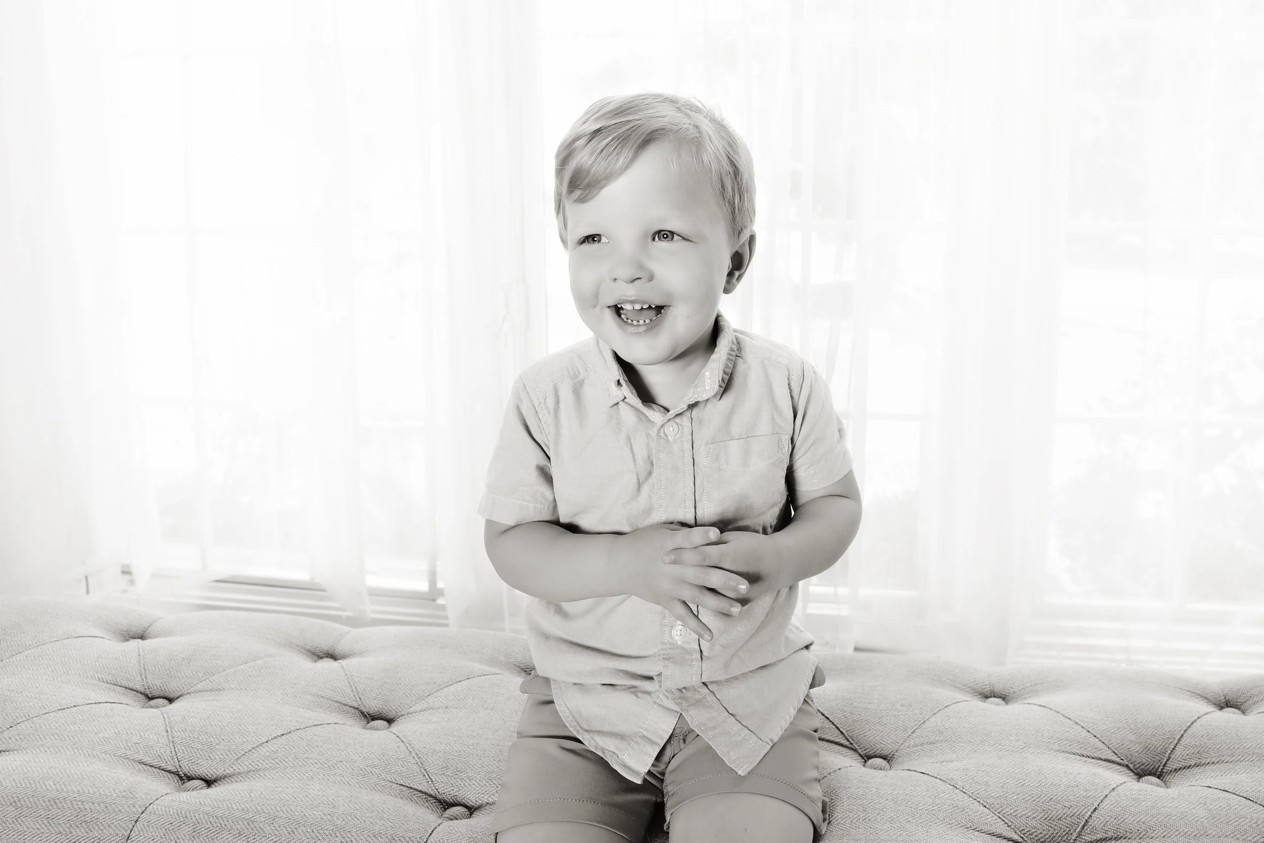 A young boy with light-colored hair, sitting on a cushioned surface in front of sheer curtains, smiling and looking to the side.