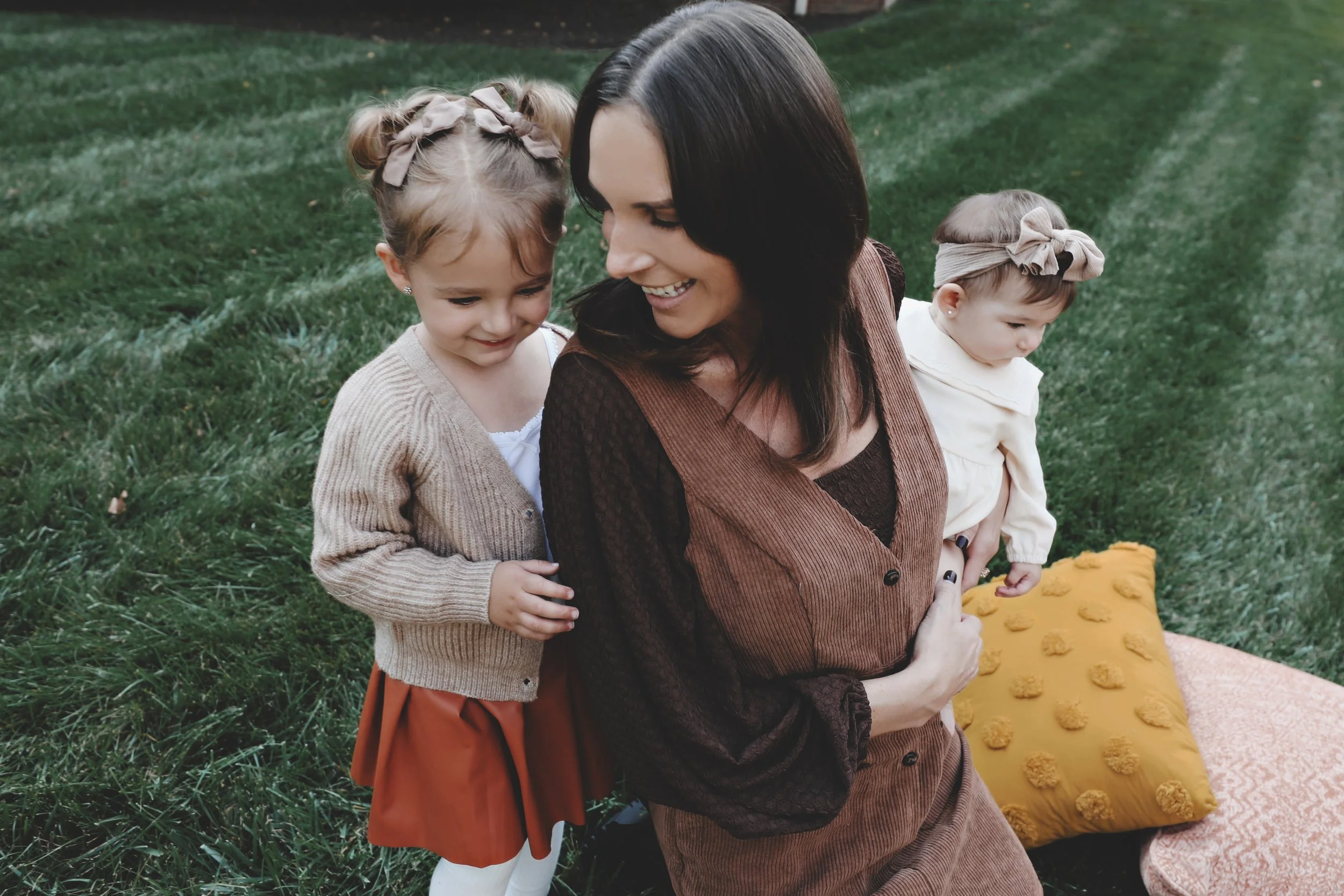 A woman outdoors on a grassy area with two young girls, one hugging her from behind and the other standing nearby, all smiling and enjoying the moment.