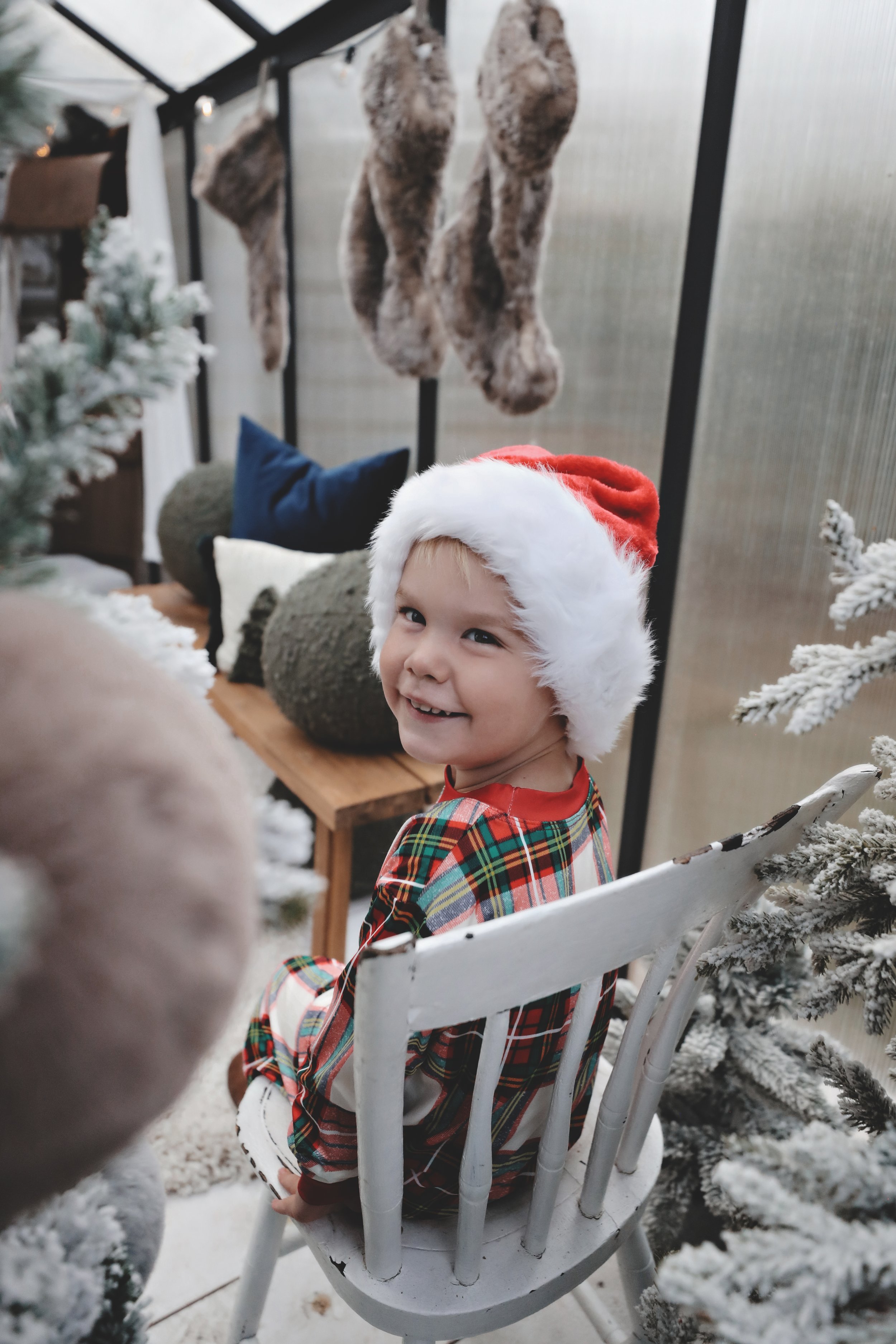 A young child wearing a red and white Santa hat and plaid pajamas, smiling and turning to look back while sitting on a white chair in a decorated winter or Christmas scene with a snow-covered Christmas tree and stockings hanging in the background.