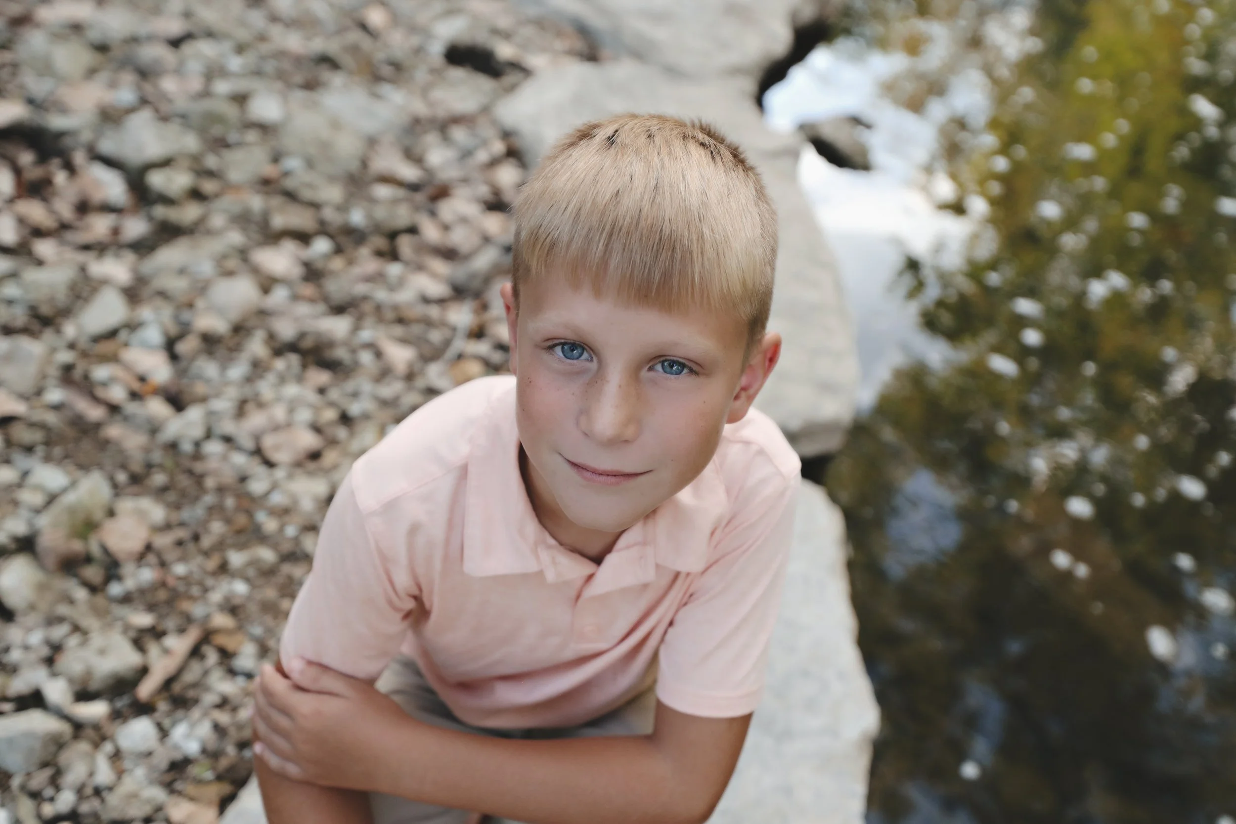 A young boy with blonde hair and blue eyes wearing a light pink shirt, leaning slightly over a rock ledge near water, looking up at the camera with a slight smile.