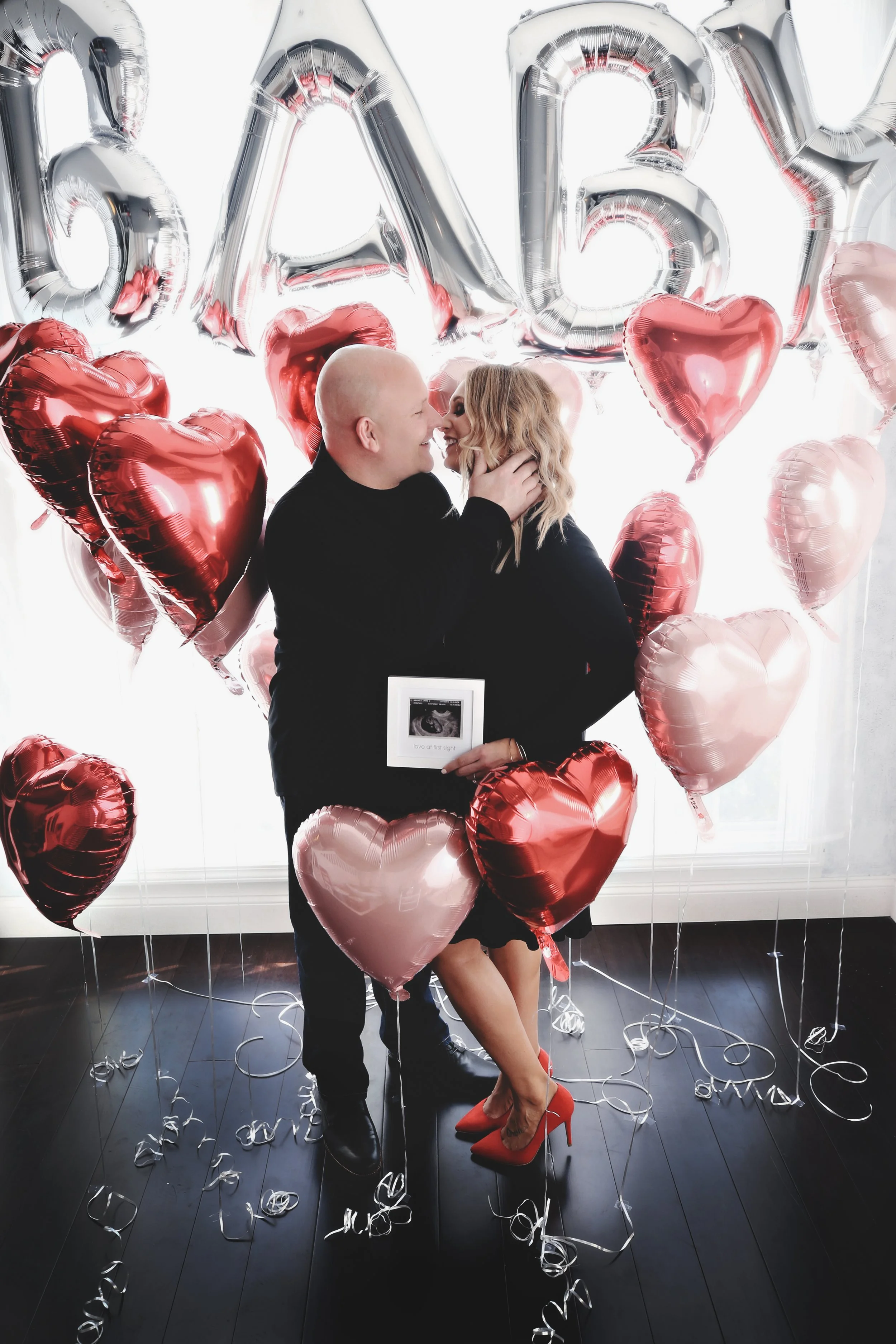 Couple standing close together, with the man holding an ultrasound picture, surrounded by red and pink heart-shaped balloons, with silver balloon letters spelling 'BABY' in the background, celebrating pregnancy.
