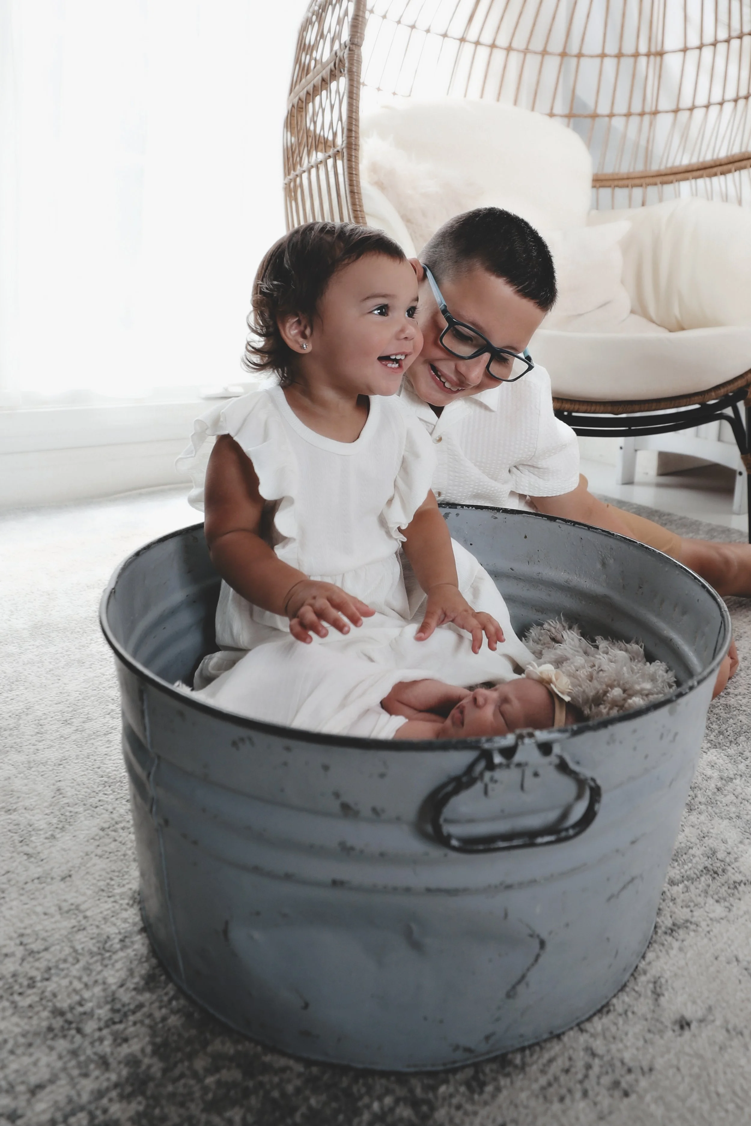 A young girl and man smiling with a baby lying in a metal tub in a bright room.