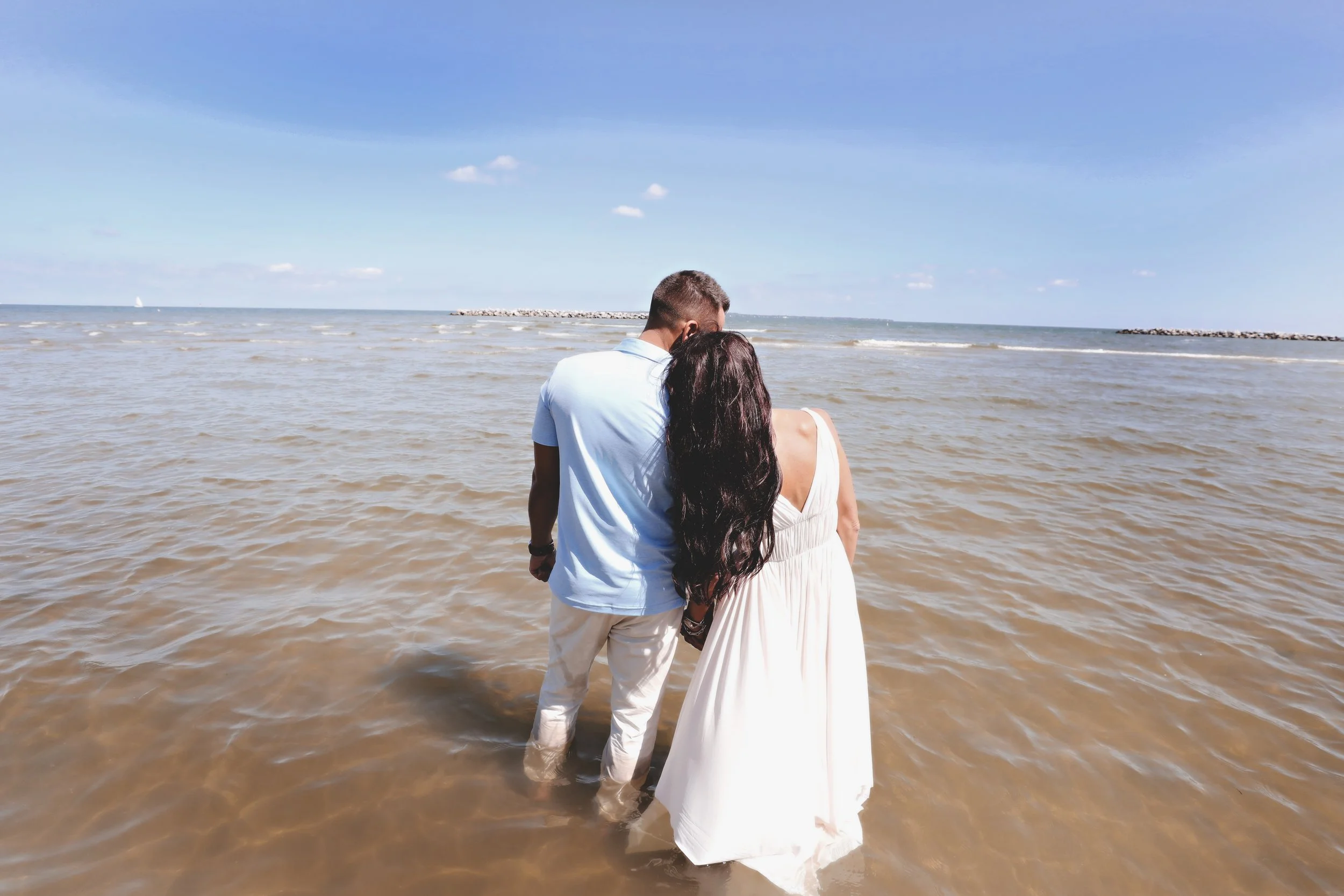 A couple holding hands and standing in the shallow water at the beach, facing the ocean on a sunny day with a partly cloudy sky.