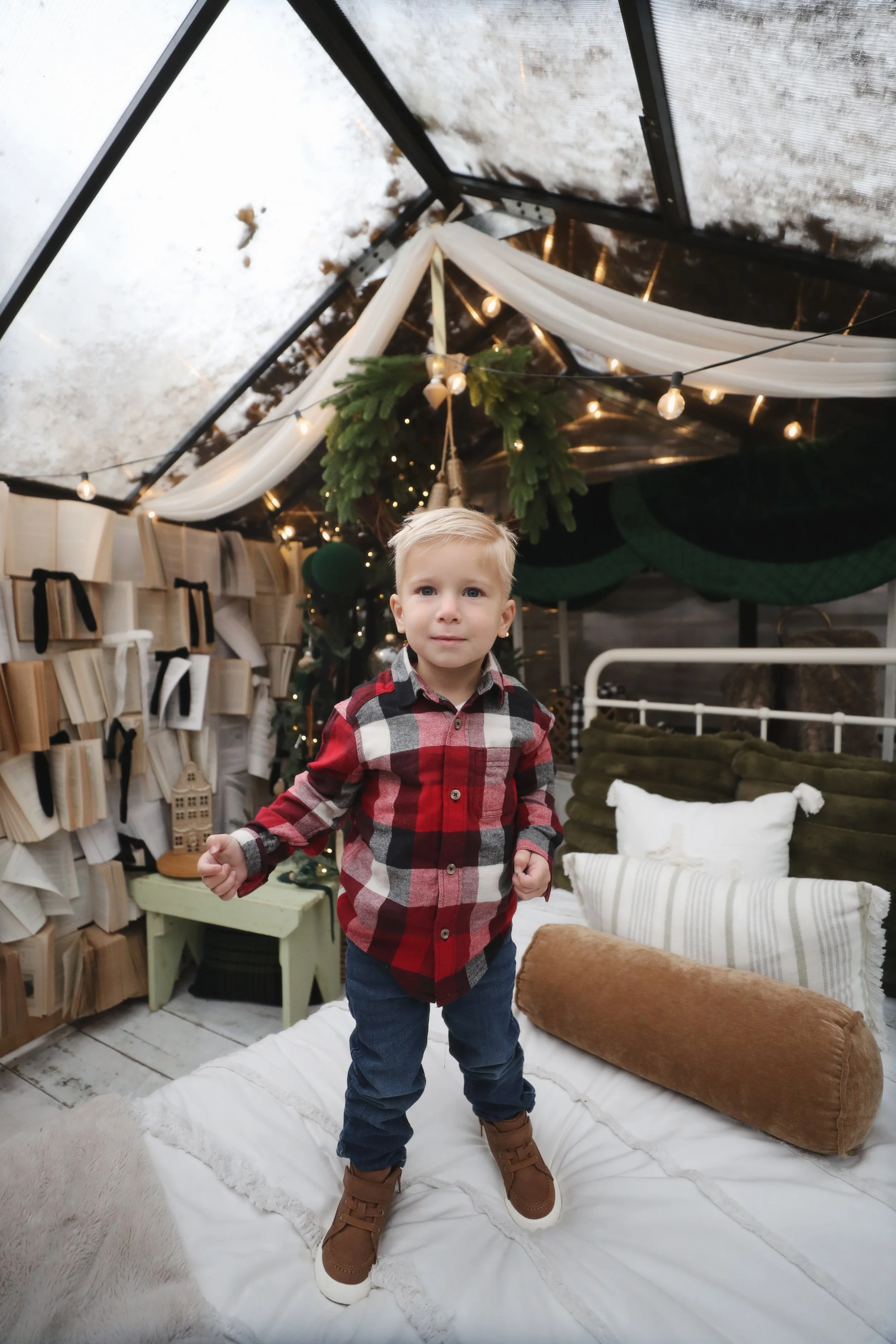 Young boy standing on a bed inside a decorated tent with Christmas greenery and fairy lights.
