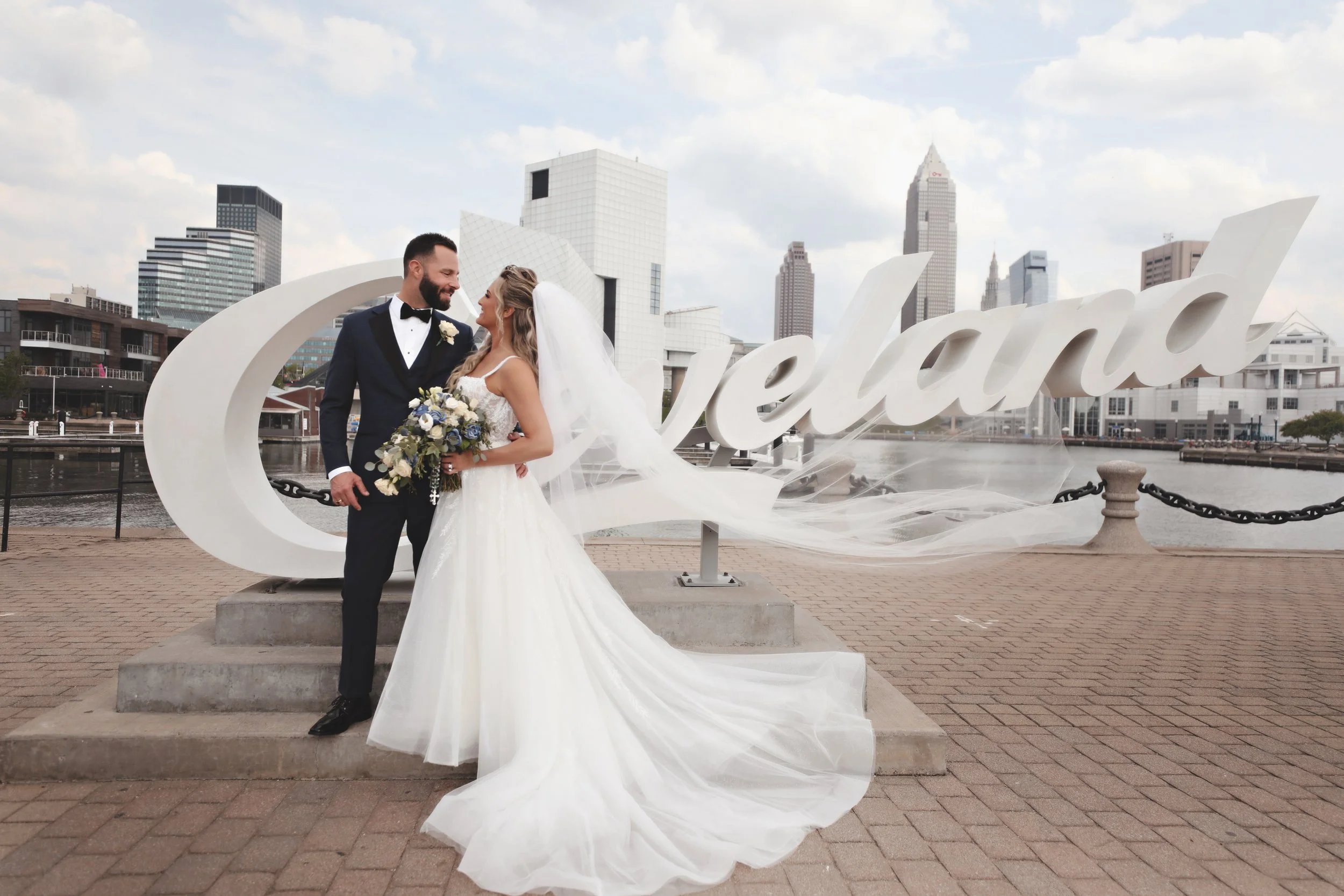 Wedding couple standing near Cleveland sign by a river, with city skyline in background.