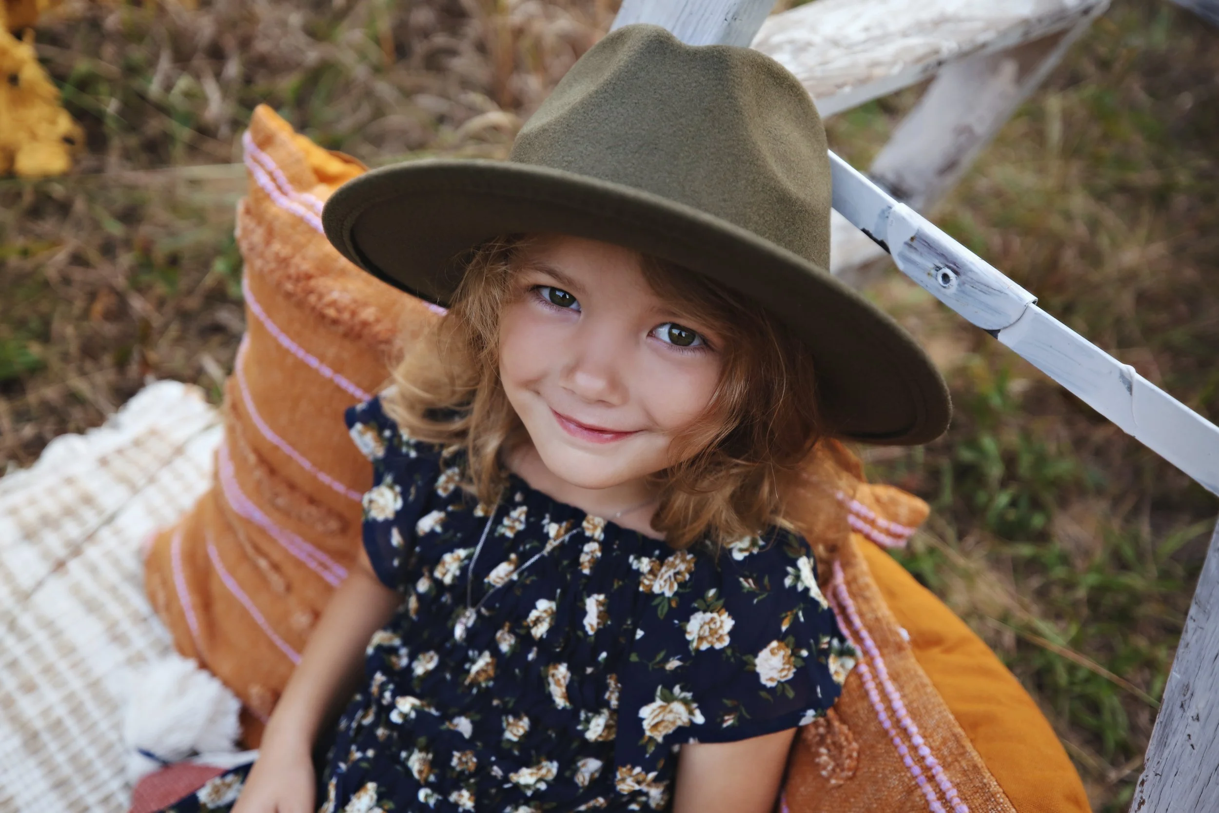 A young girl with curly hair wearing a wide-brimmed hat and a floral dress, sitting on a blanket outdoors, smiling up at the camera.