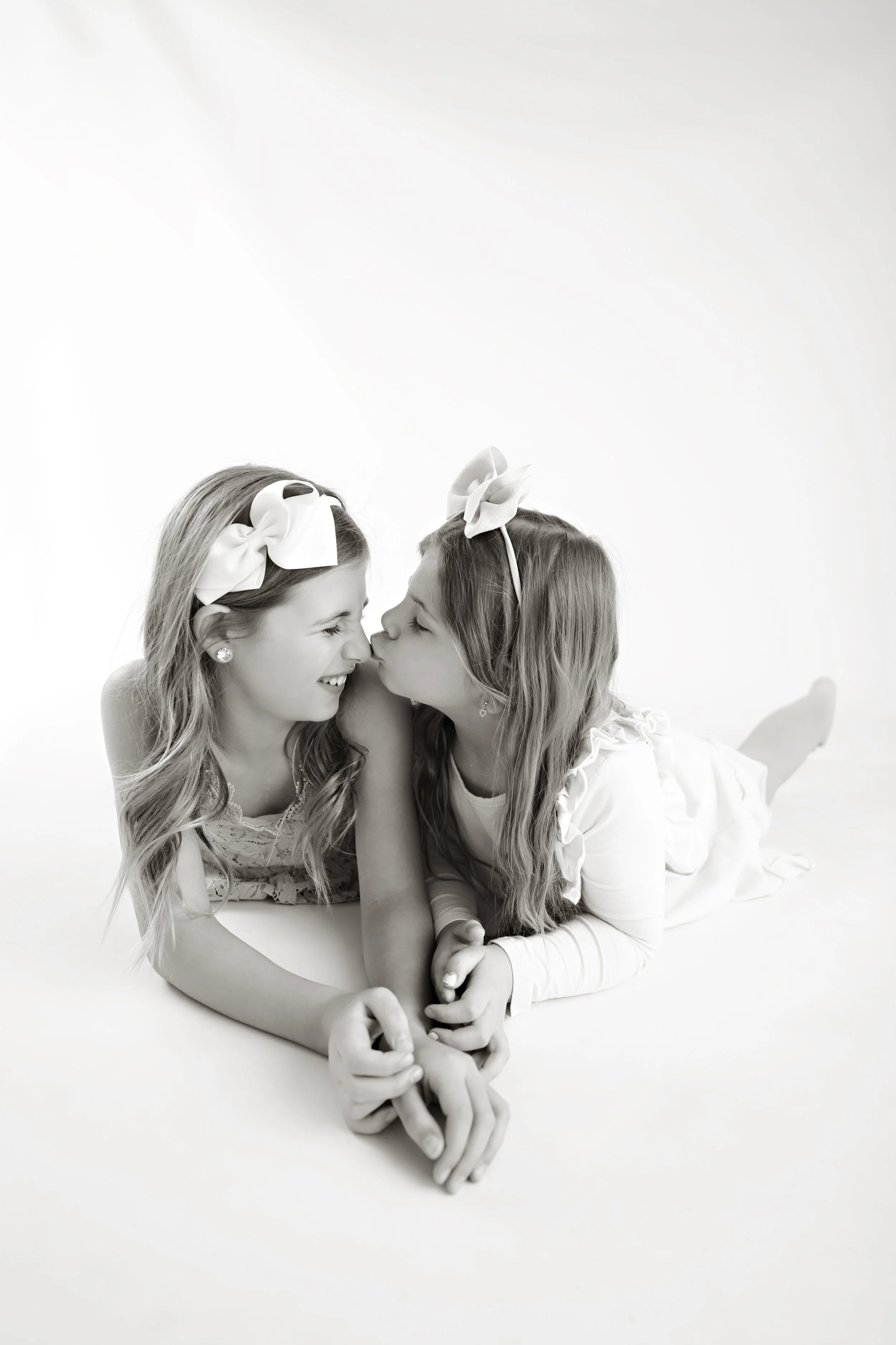 Two young girls lying on the floor, face to face, touching noses, smiling and holding hands. They are wearing headbands with bows and light-colored dresses, in a bright, minimal background.