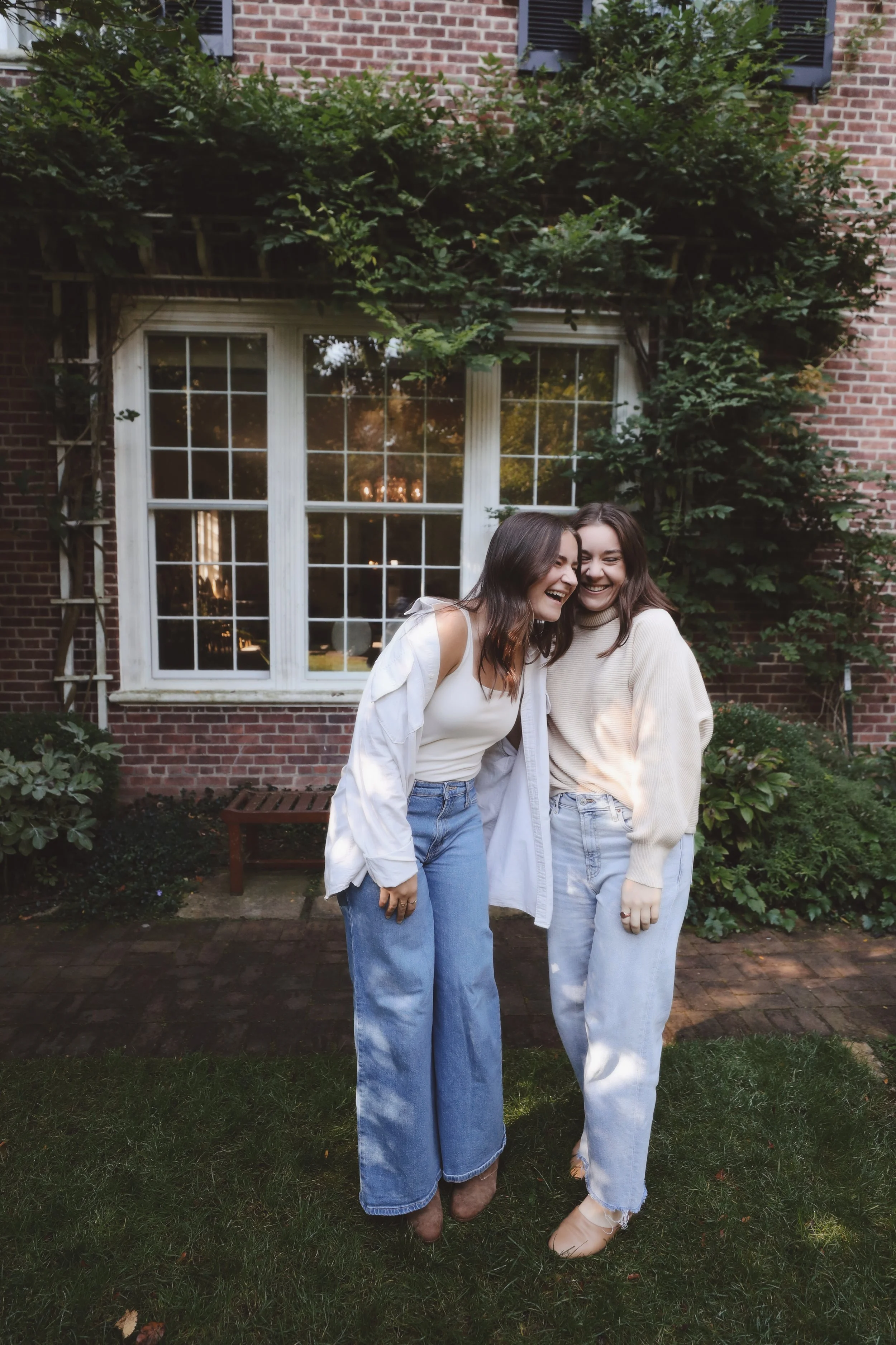 Two women with dark hair laughing and leaning on each other, standing outside in front of a red brick house with a large window and greenery.
