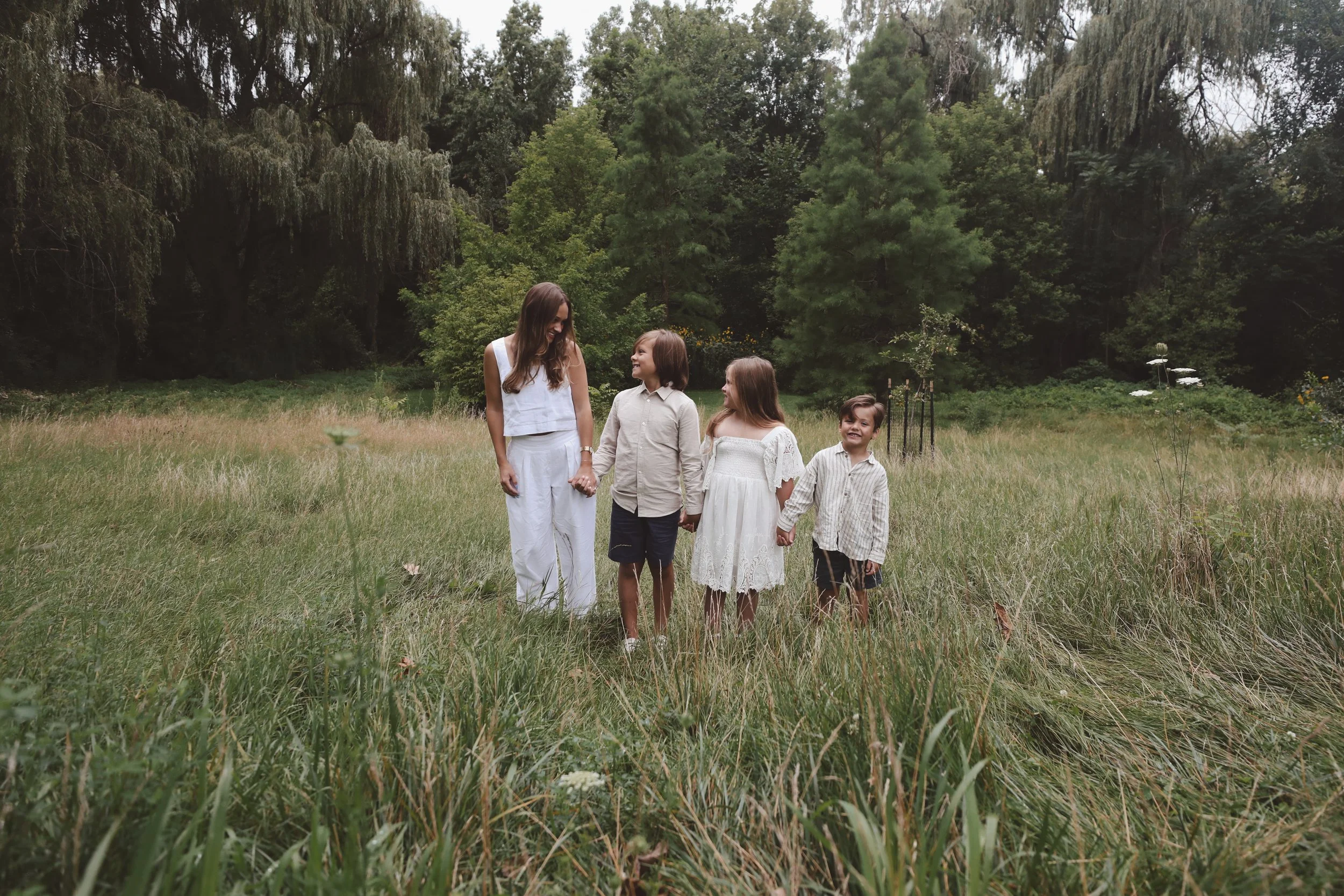 A woman and three children walking hand-in-hand in a grassy field with trees in the background on a cloudy day.