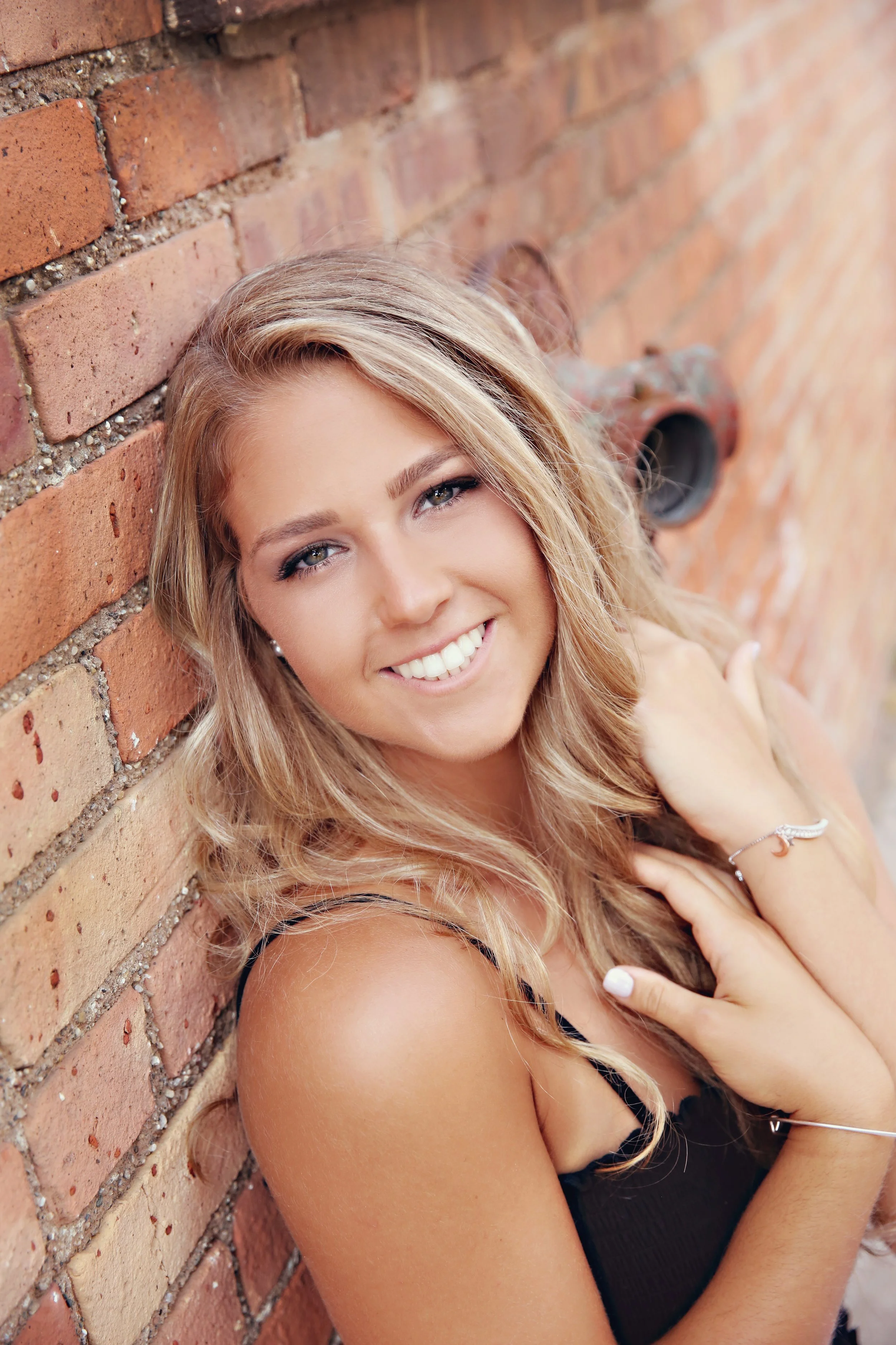 A young woman with long blonde hair, smiling, leaning against a brick wall, wearing a black top.