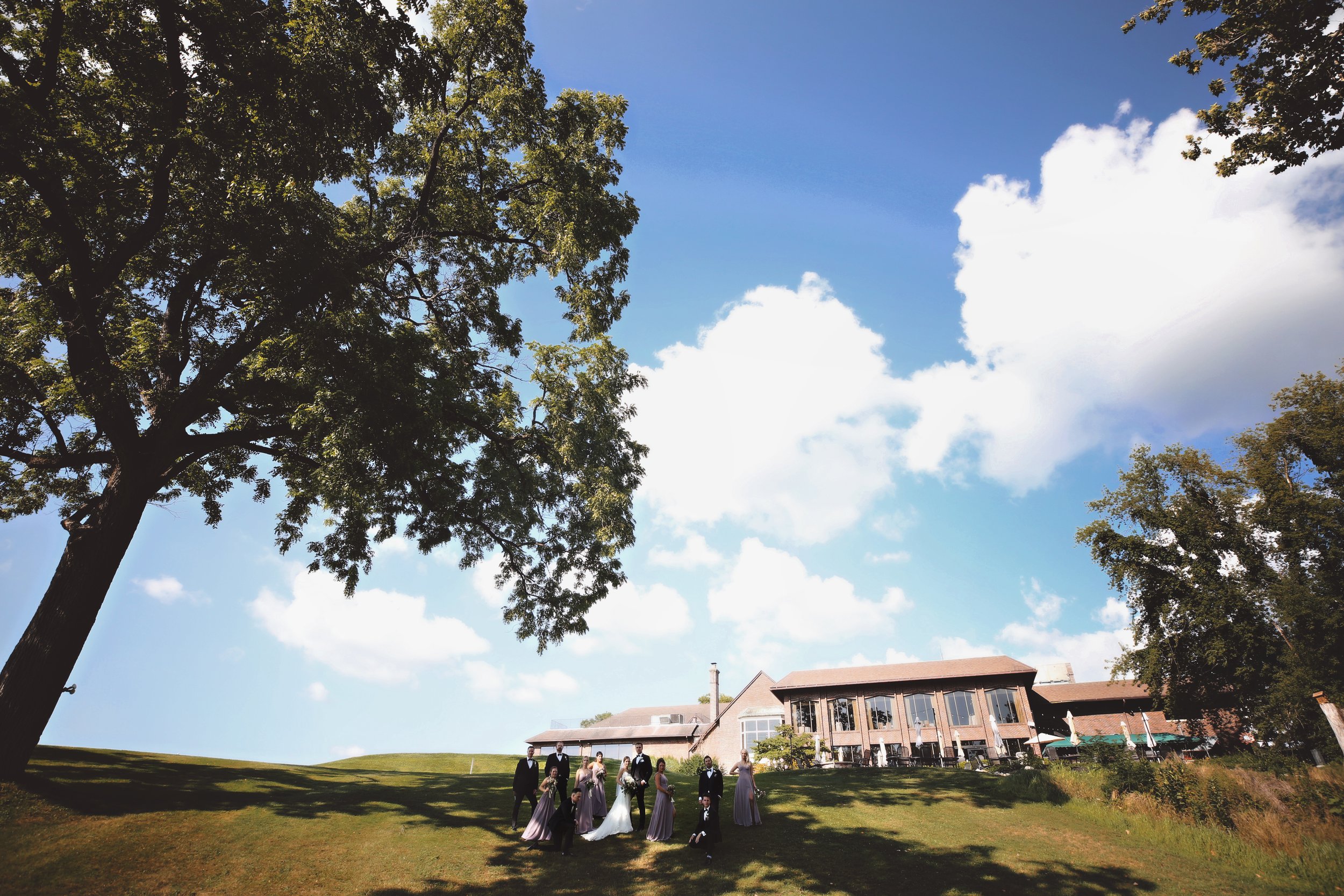 A group of people dressed in formal attire standing outside a house on a grassy lawn with large trees and a blue sky with some clouds.