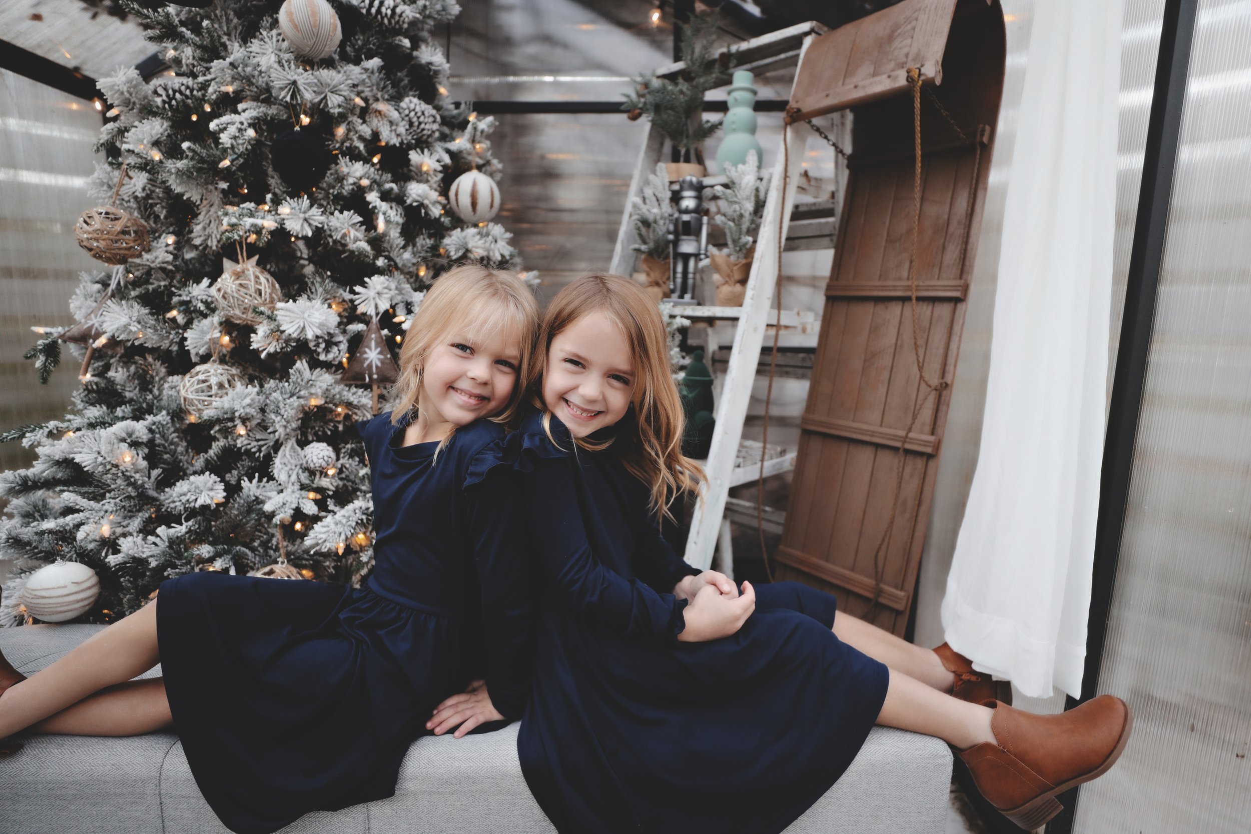 Two young girls with blonde and brunette hair, smiling, sitting on a gray bench in front of a decorated Christmas tree with ornaments and lights, in a cozy indoor space with holiday decor.