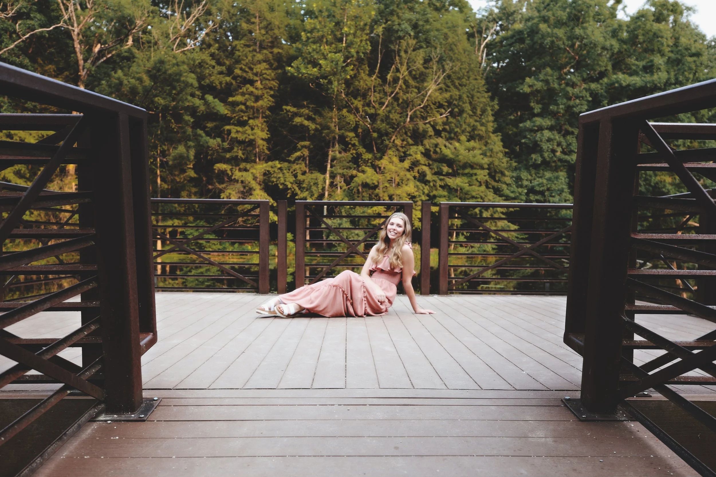 A young woman in a pink dress sitting on a wooden deck with a forest in the background, smiling at the camera.