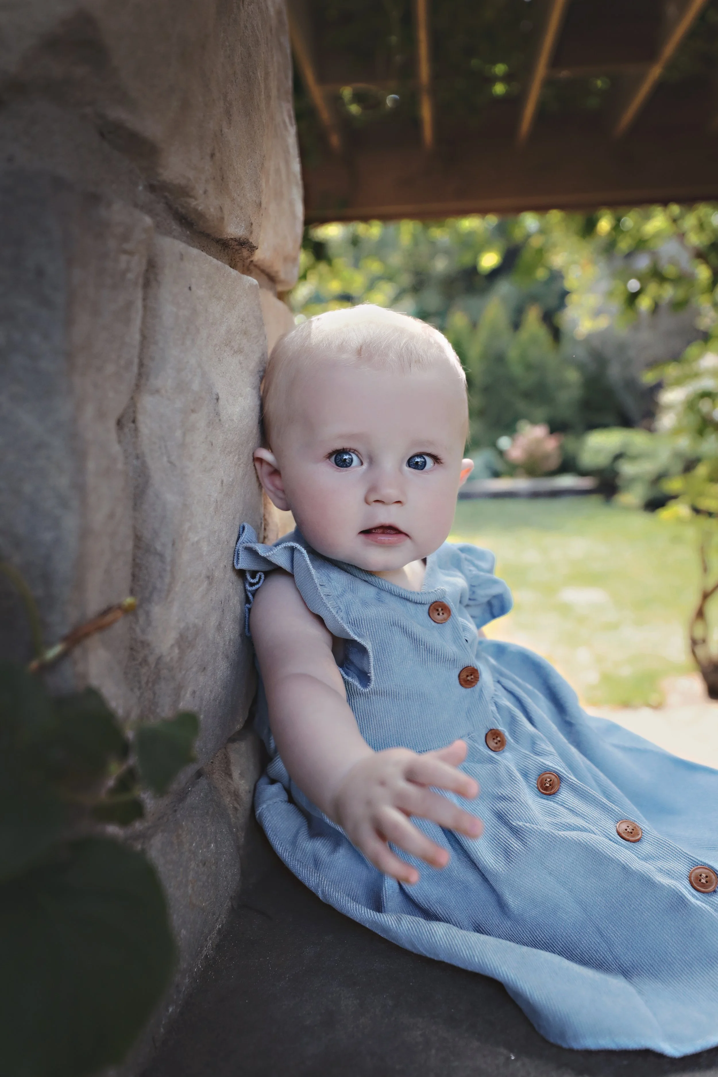 Young child with blue eyes and blonde hair, wearing a blue dress with buttons, sitting outdoors next to a stone wall with green trees in the background.
