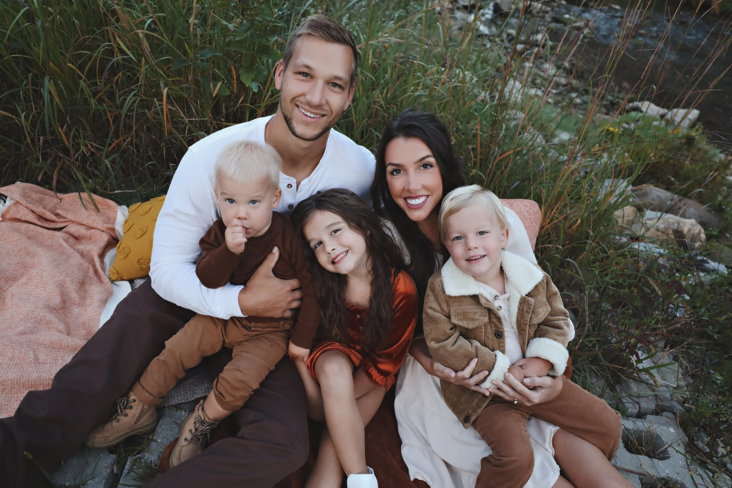 Family of six sitting outdoors on a blanket, surrounded by tall grass and rocks, smiling at the camera.