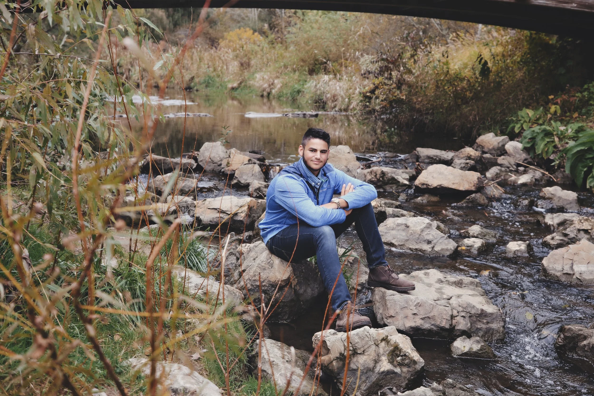 A young man sitting on rocks by a creek in a natural, wooded area during autumn with colorful foliage.