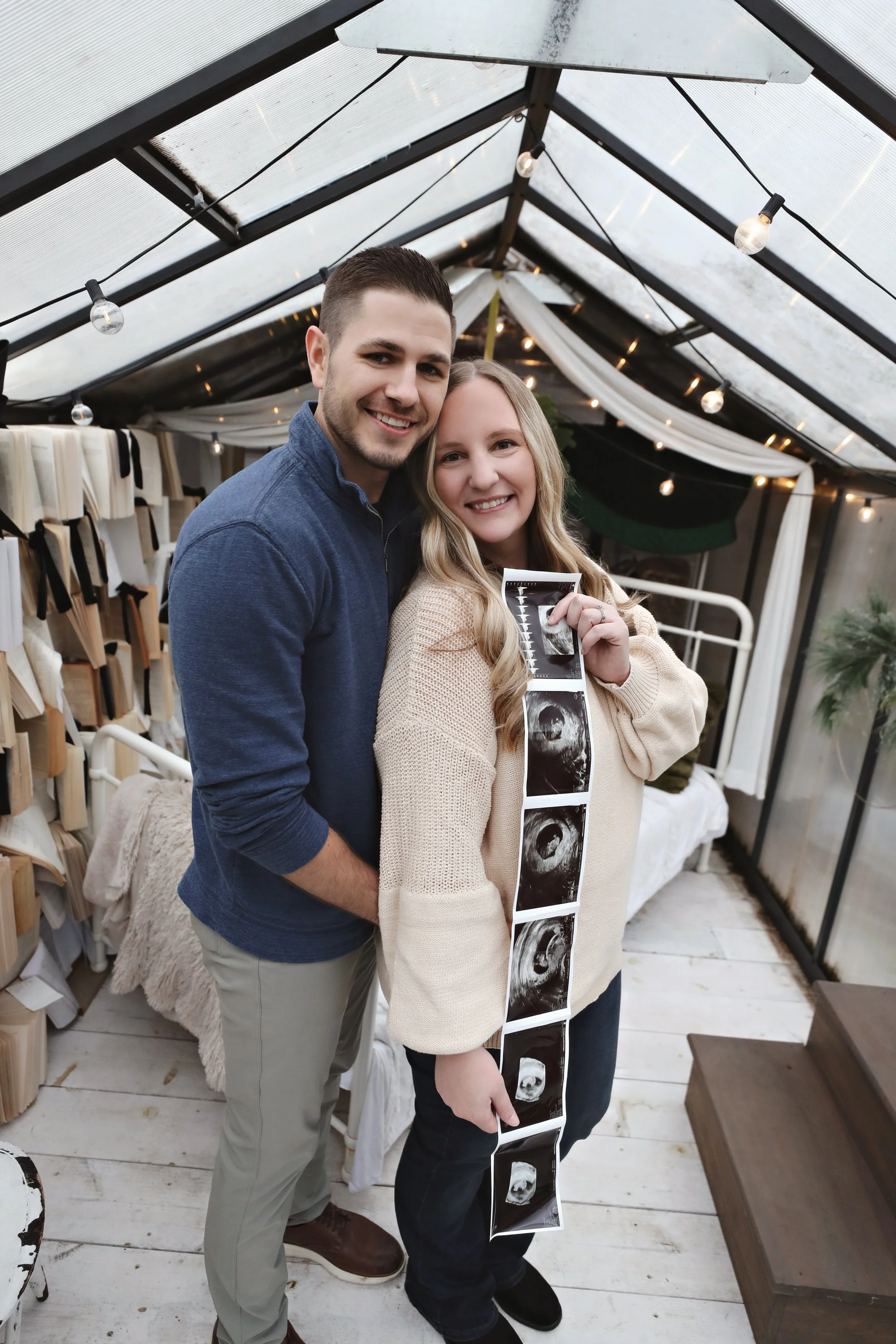 A couple, woman and man, smiling inside a greenhouse, with the woman holding an ultrasound strip showing four ultrasound images.