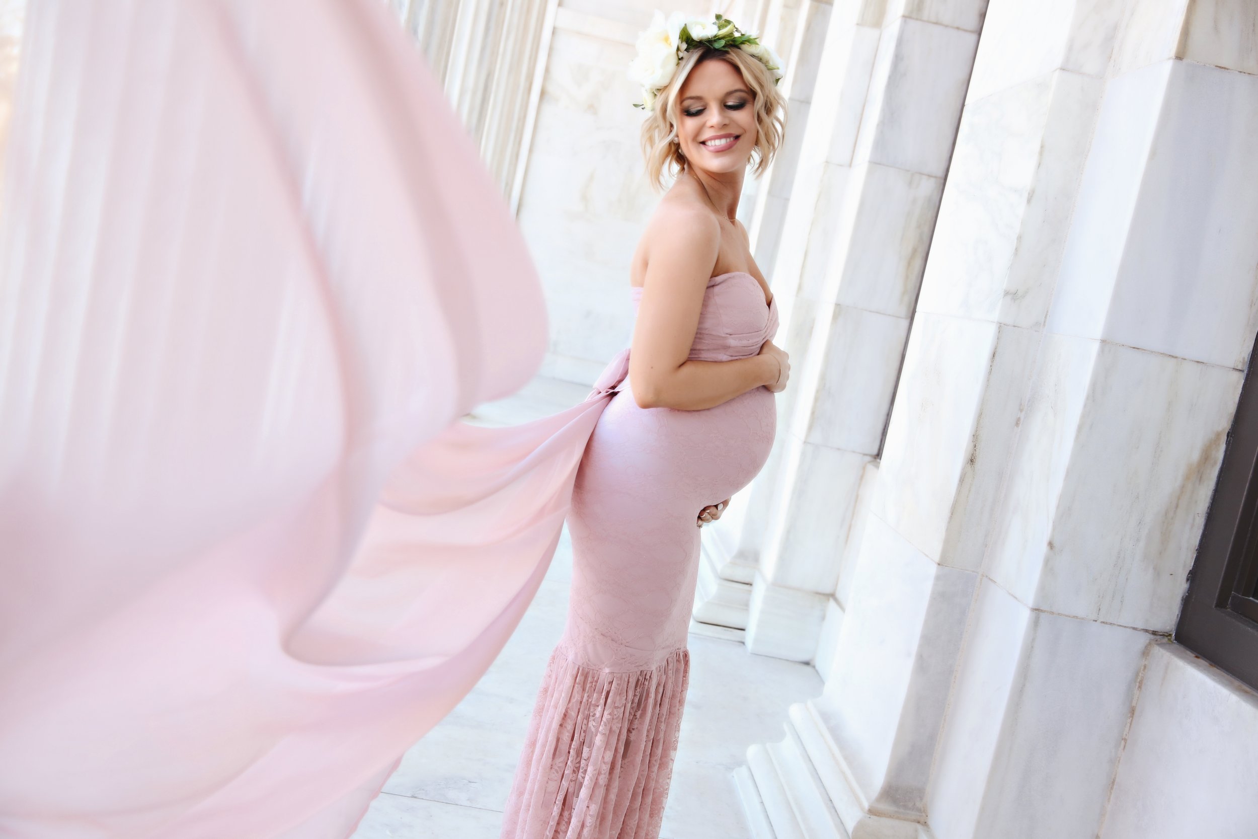 Pregnant woman in a pink dress with a floral headband smiling and holding her belly, standing in a marble corridor.