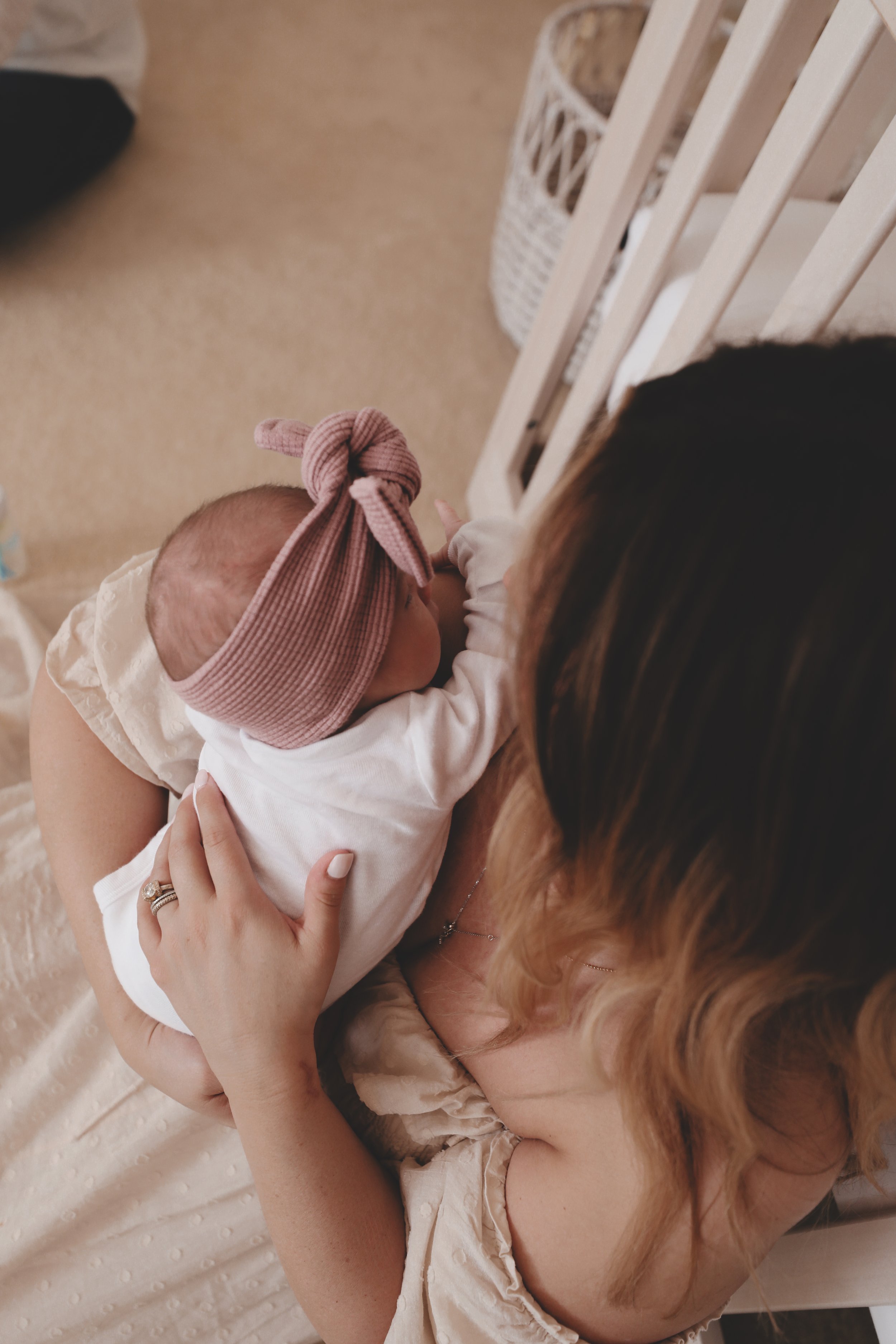 A woman holding a newborn baby on her chest, seen from above. The baby is wearing a pink headband with a bow and a white outfit. The setting appears to be a nursery with a crib and a wicker basket.