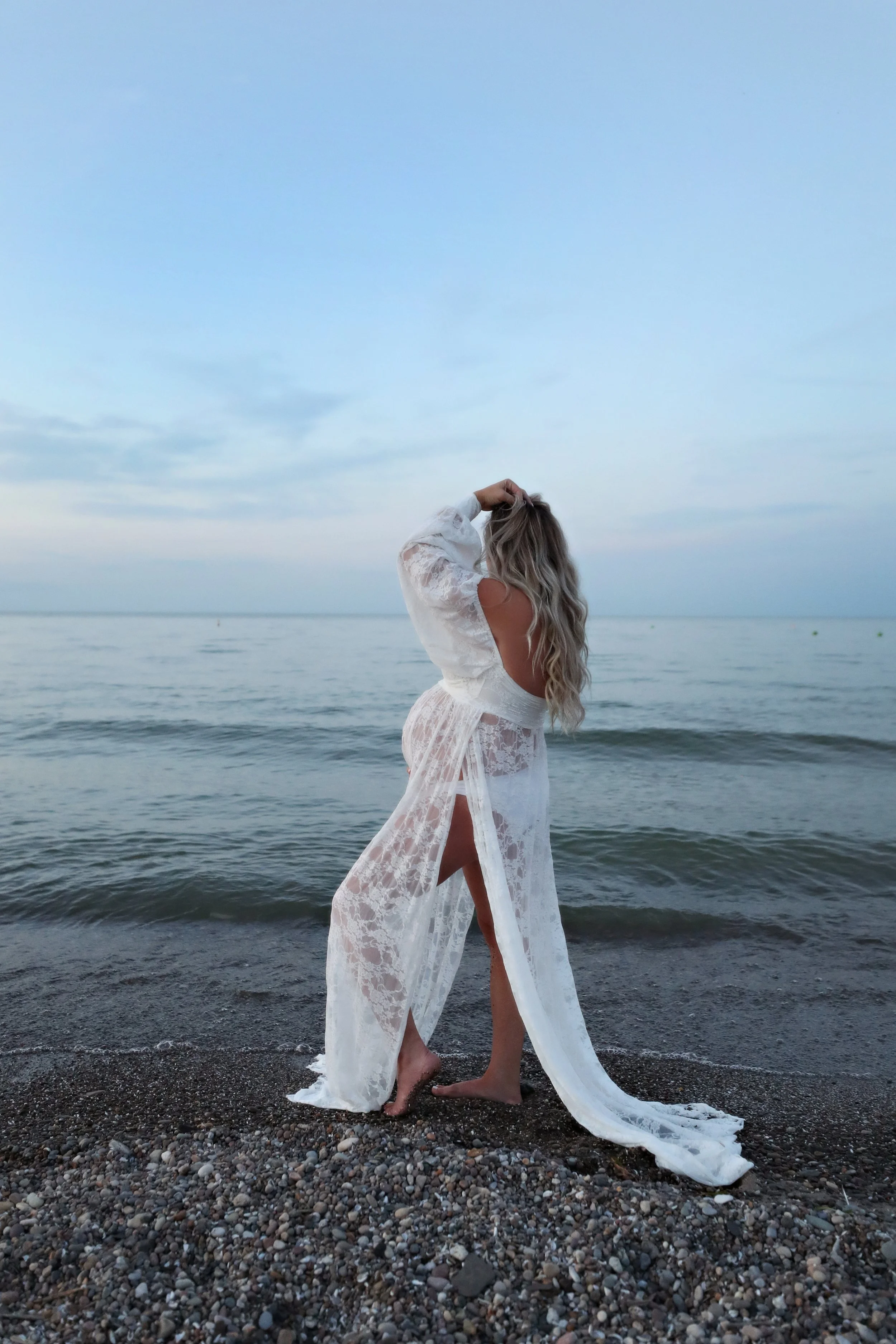 A woman in a white lace dress standing on a pebbled beach by the ocean with her back to the camera, touching her hair, under a cloudy sky.