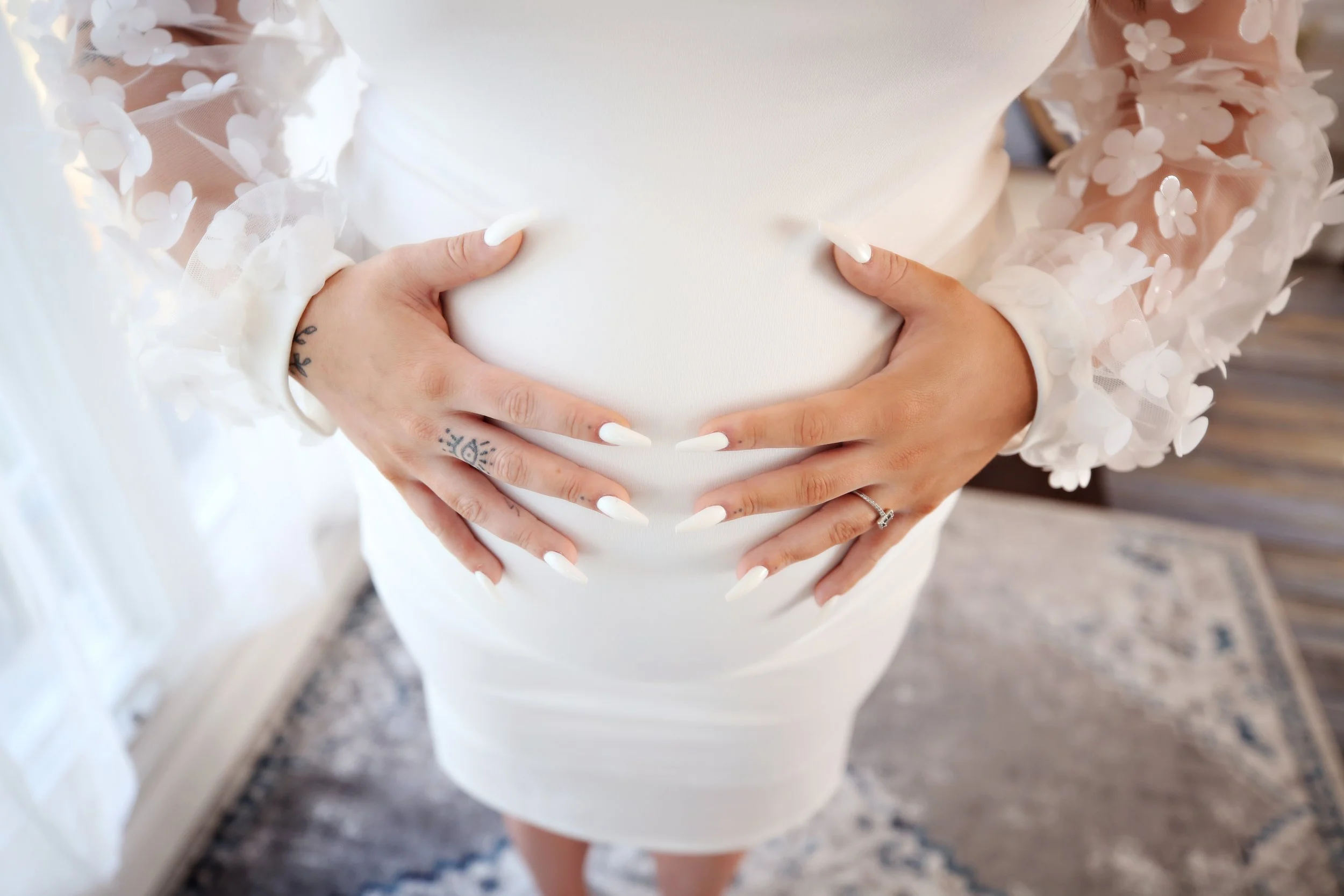 Pregnant woman wearing a white dress with sheer sleeves decorated with floral appliqués, holding her belly with both hands adorned with rings and tattoos.