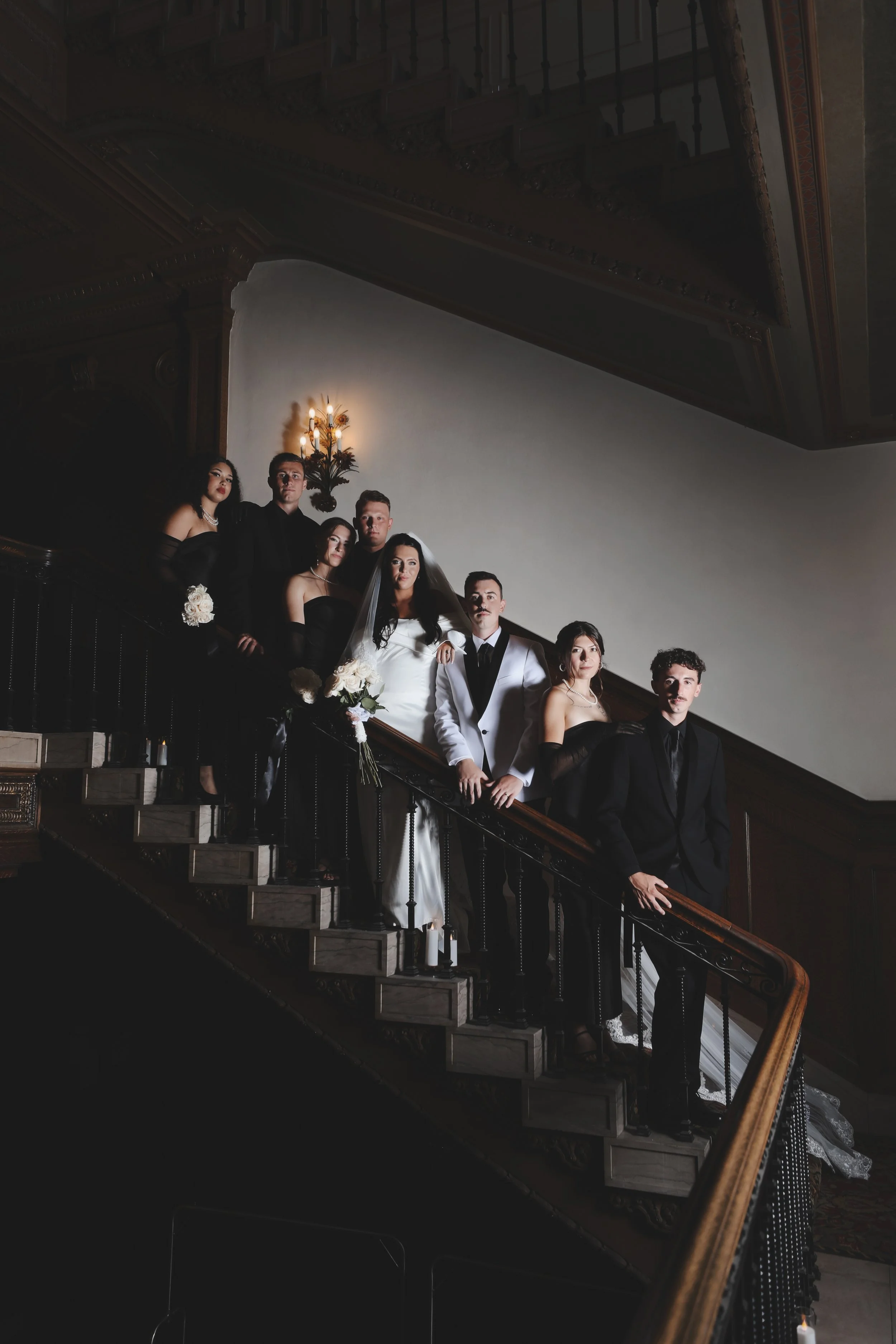 A wedding party standing on a staircase inside an elegant building, with the bride in a white gown holding a bouquet, and the groom in a tuxedo, surrounded by bridesmaids and groomsmen dressed in black and white.