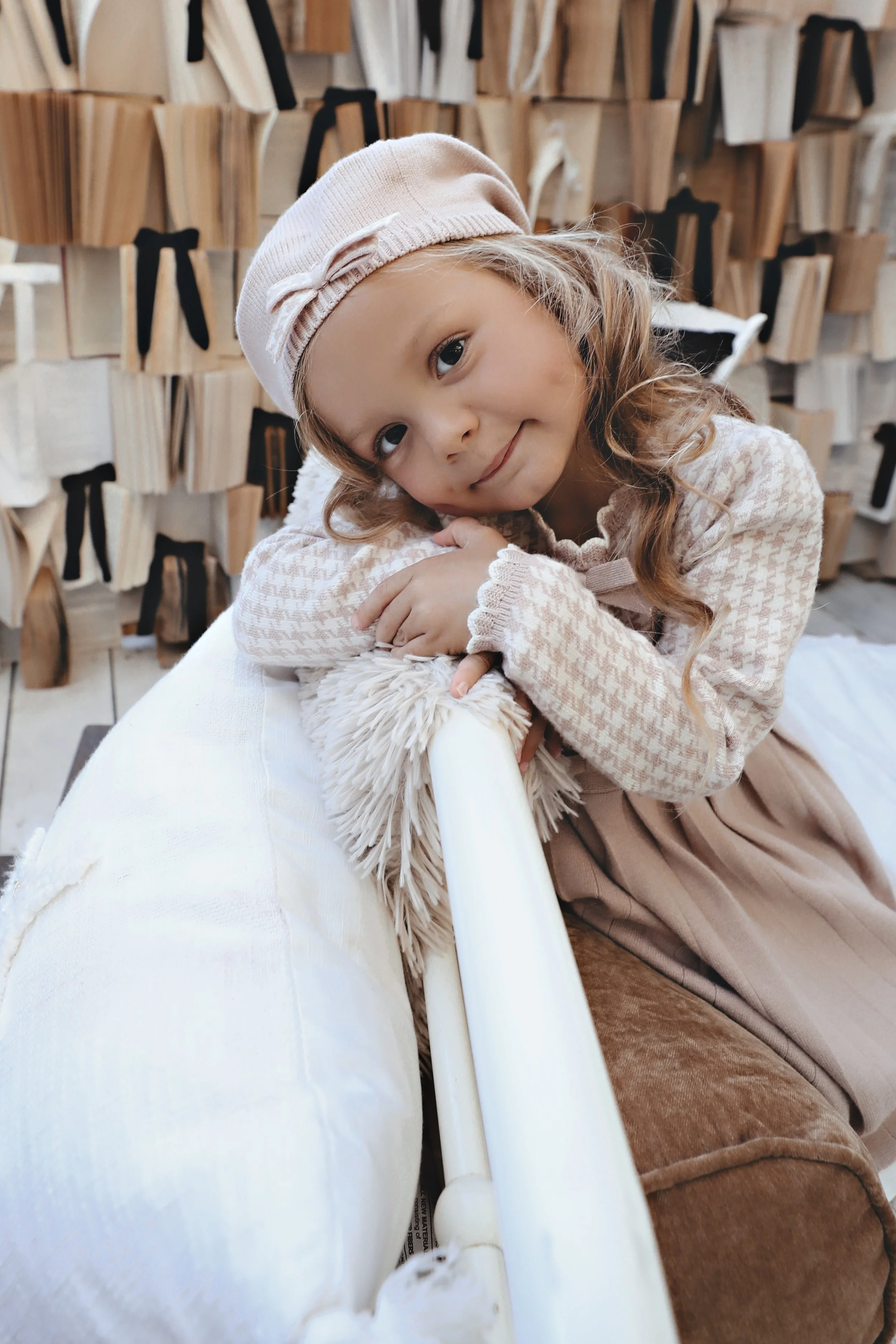A young girl with blonde hair, wearing a beige hat and patterned sweater, leaning on a white cushion, smiling, with a background of wooden books or decorative items.