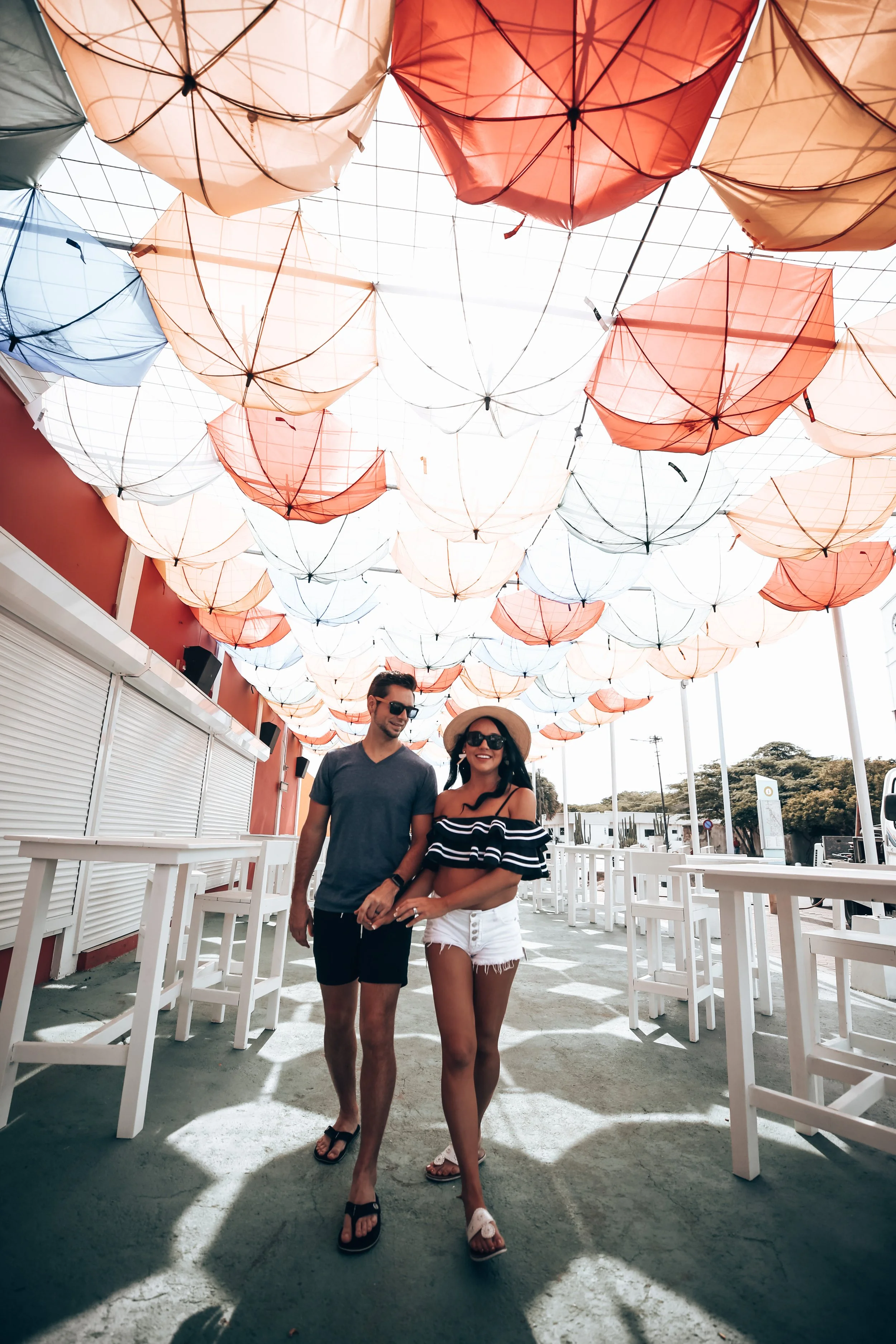 Two people, a man and a woman, walking together outdoors under umbrella-shaped decorations hanging above. The man is wearing sunglasses, a dark T-shirt, and shorts, and the woman is wearing a wide-brimmed hat, sunglasses, a striped off-shoulder top, 