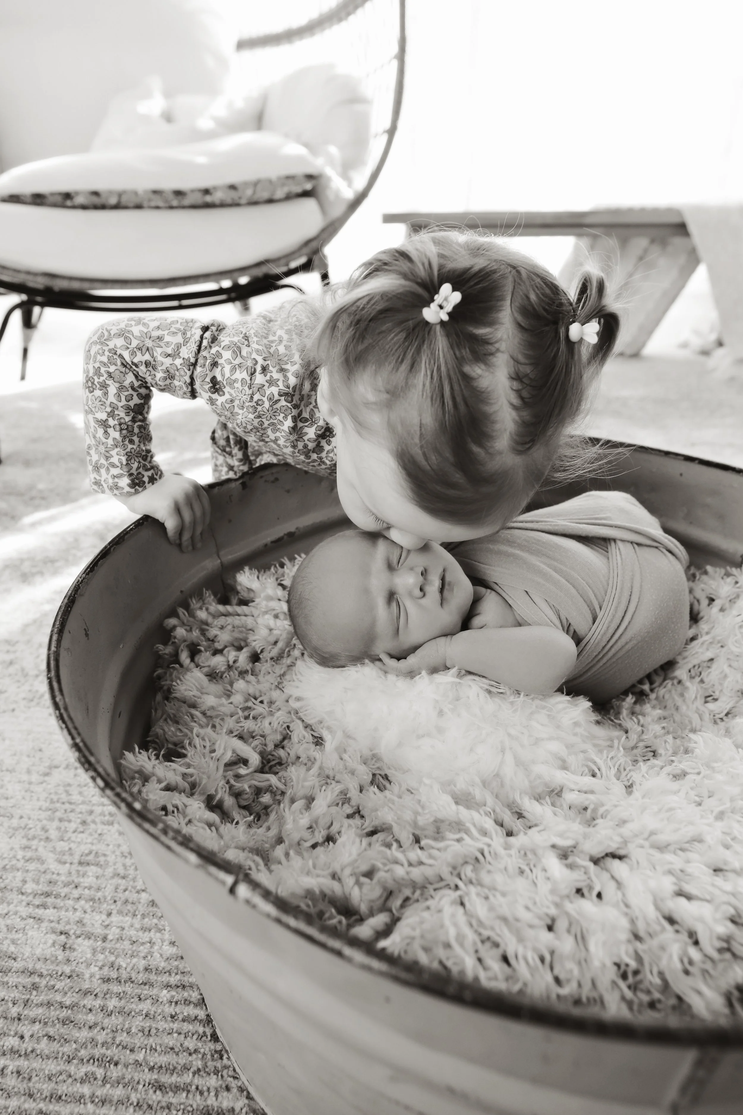 A young girl kisses a newborn baby in a cozy, furry blanket inside a wooden tub.