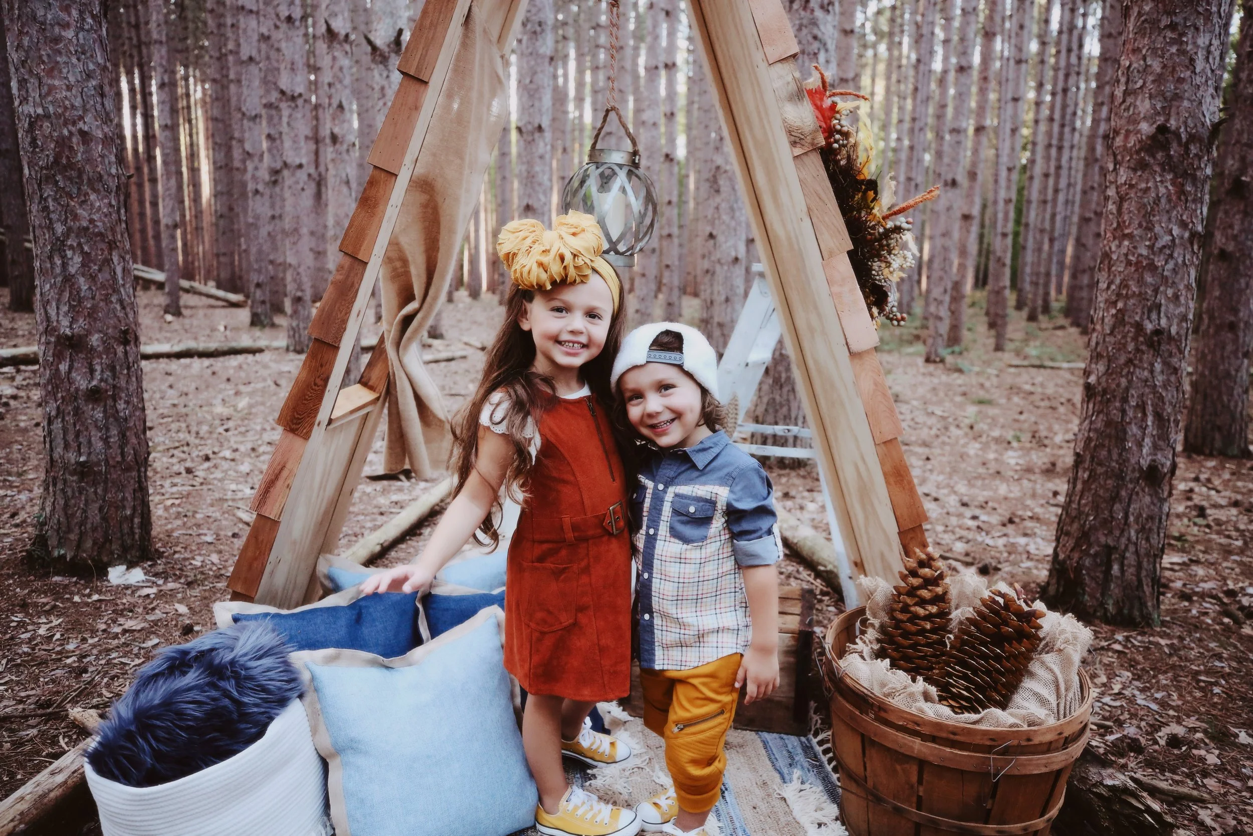 Two young children, a girl and a boy, smiling and standing together in a forest, in front of a wooden teepee decorated with autumn-themed items, including a lantern, a blanket, pillows, and pinecones.