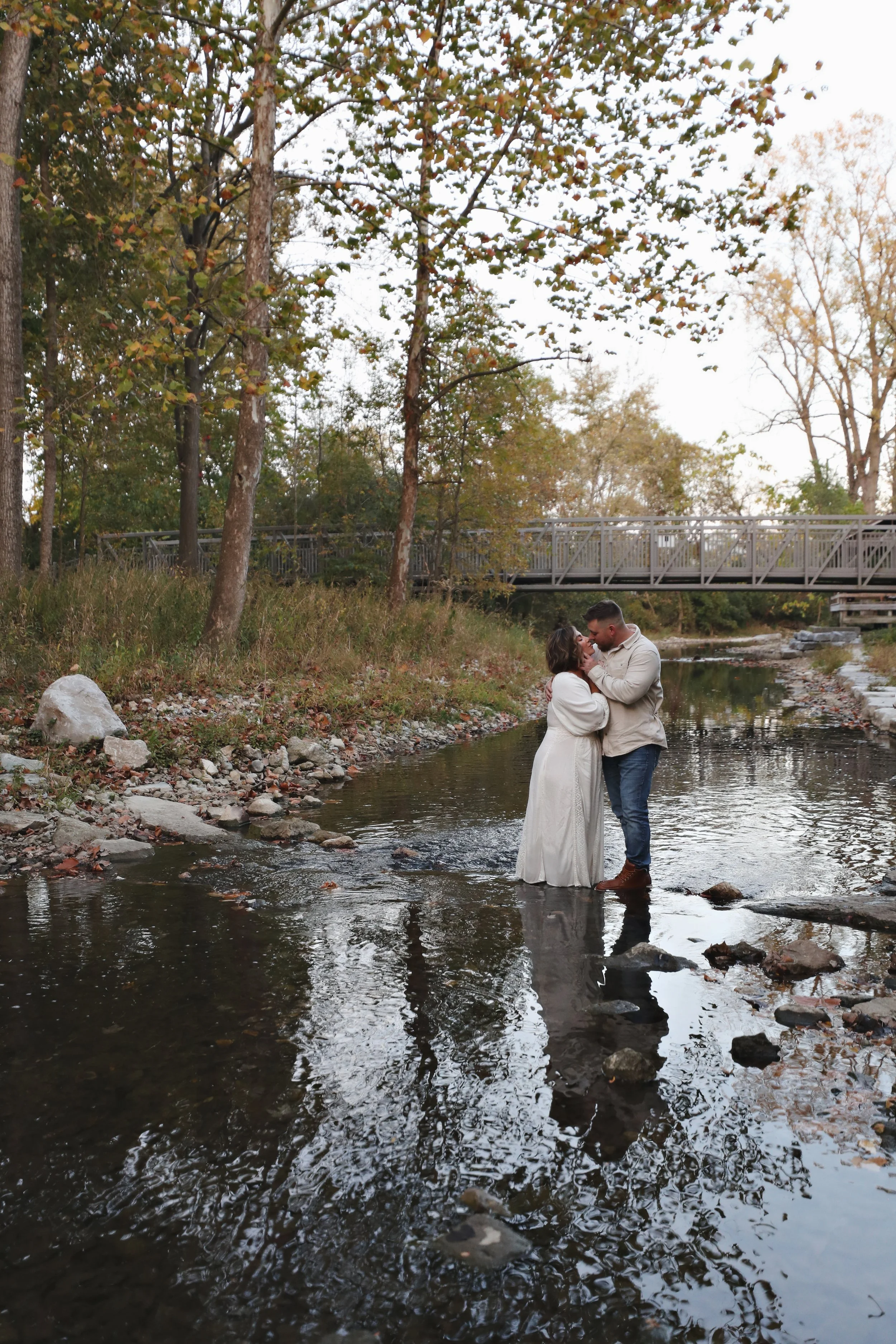 A couple kissing in a shallow river surrounded by trees with fall foliage, with a bridge in the background.