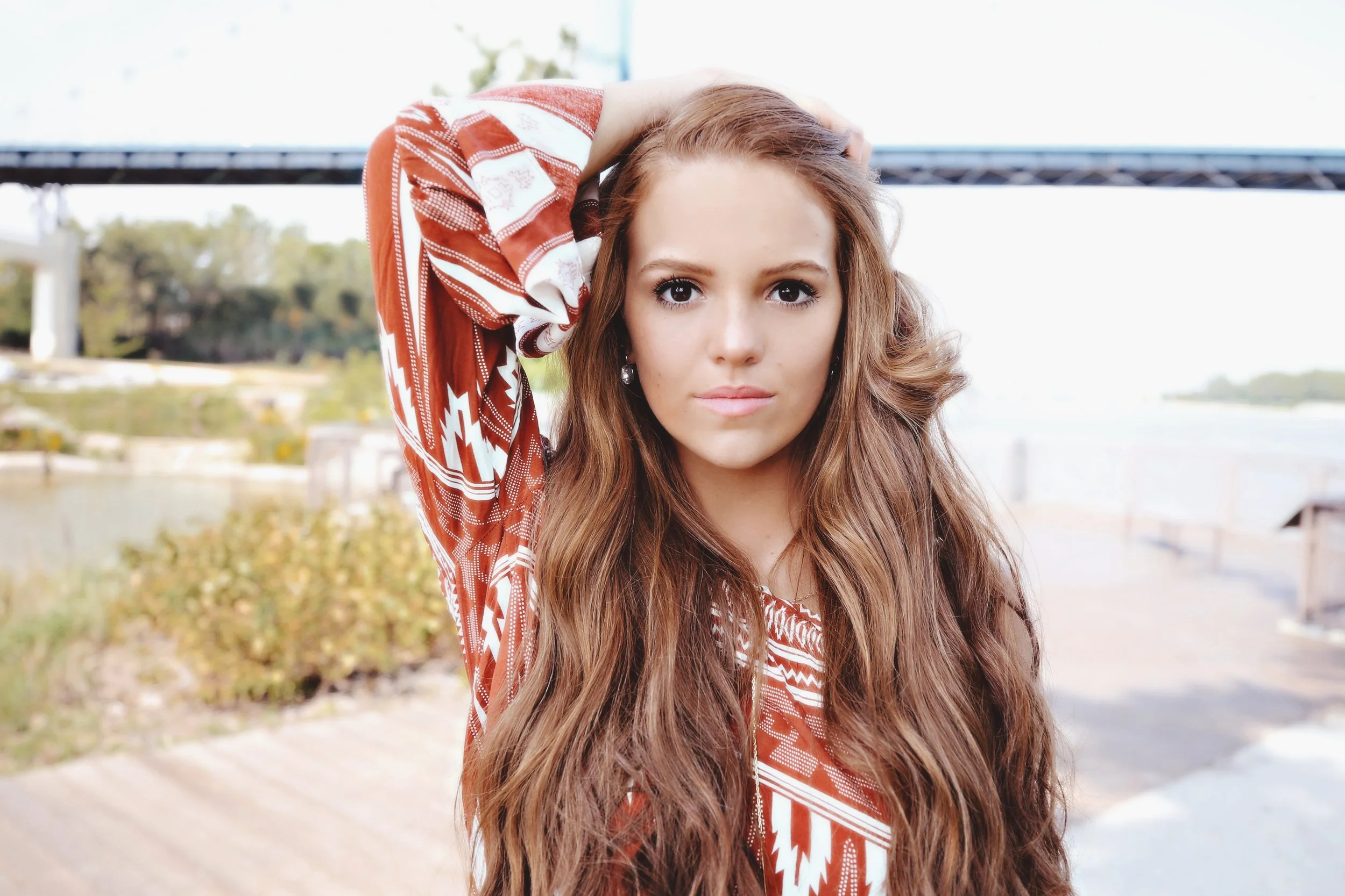 A young woman with long wavy brown hair, wearing a red patterned top, standing outdoors near a river or lake with a bridge in the background.