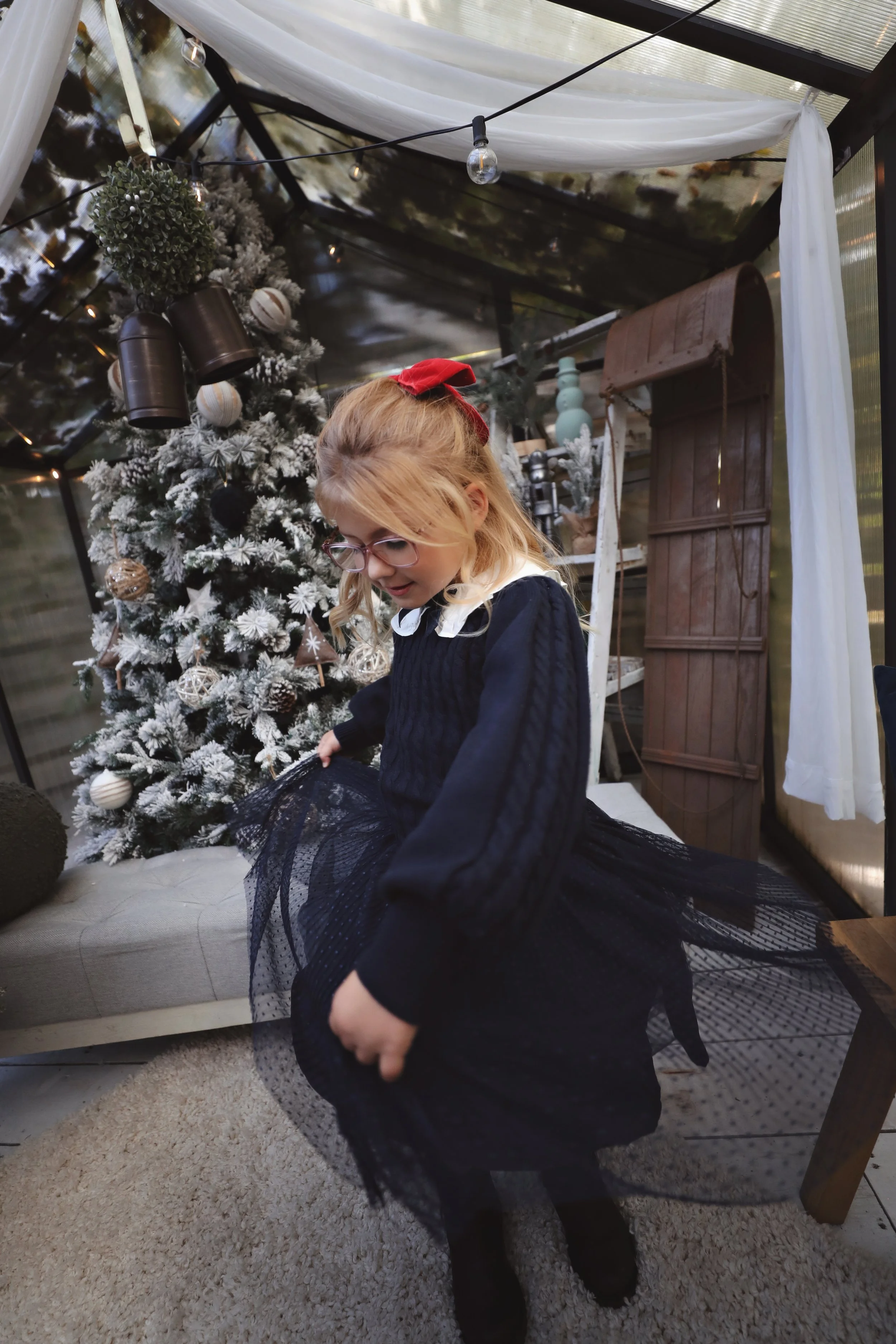 A young girl with glasses and a red bow in her hair is dressed in a black dress with a tulle skirt, standing inside a decorated Christmas-themed space with a frosted Christmas tree and holiday ornaments.