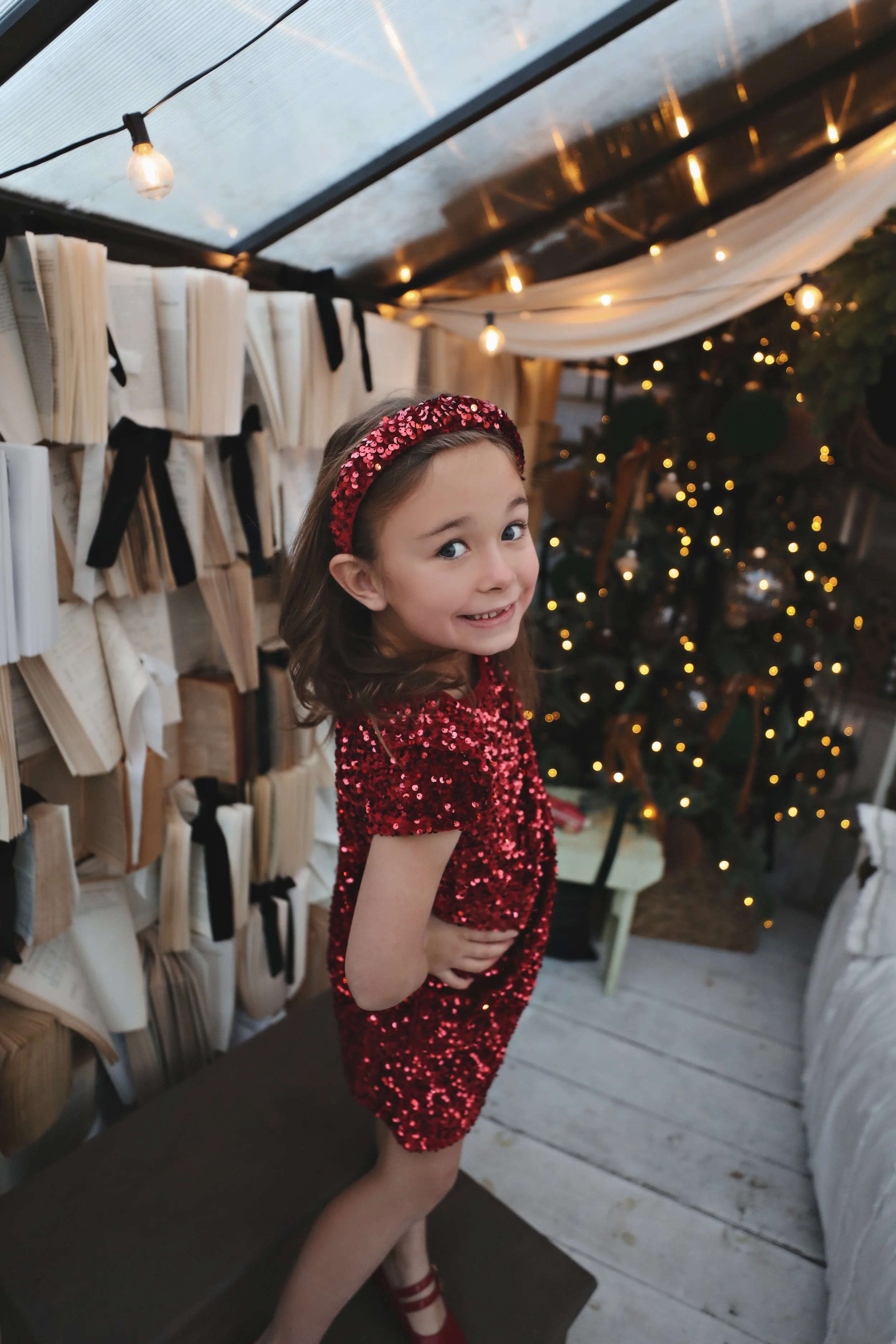 Young girl in a red sequined dress and matching headband smiling and posing indoors near a decorated Christmas tree with lights, with books stacked along the wall and a string of lights hanging from the ceiling.
