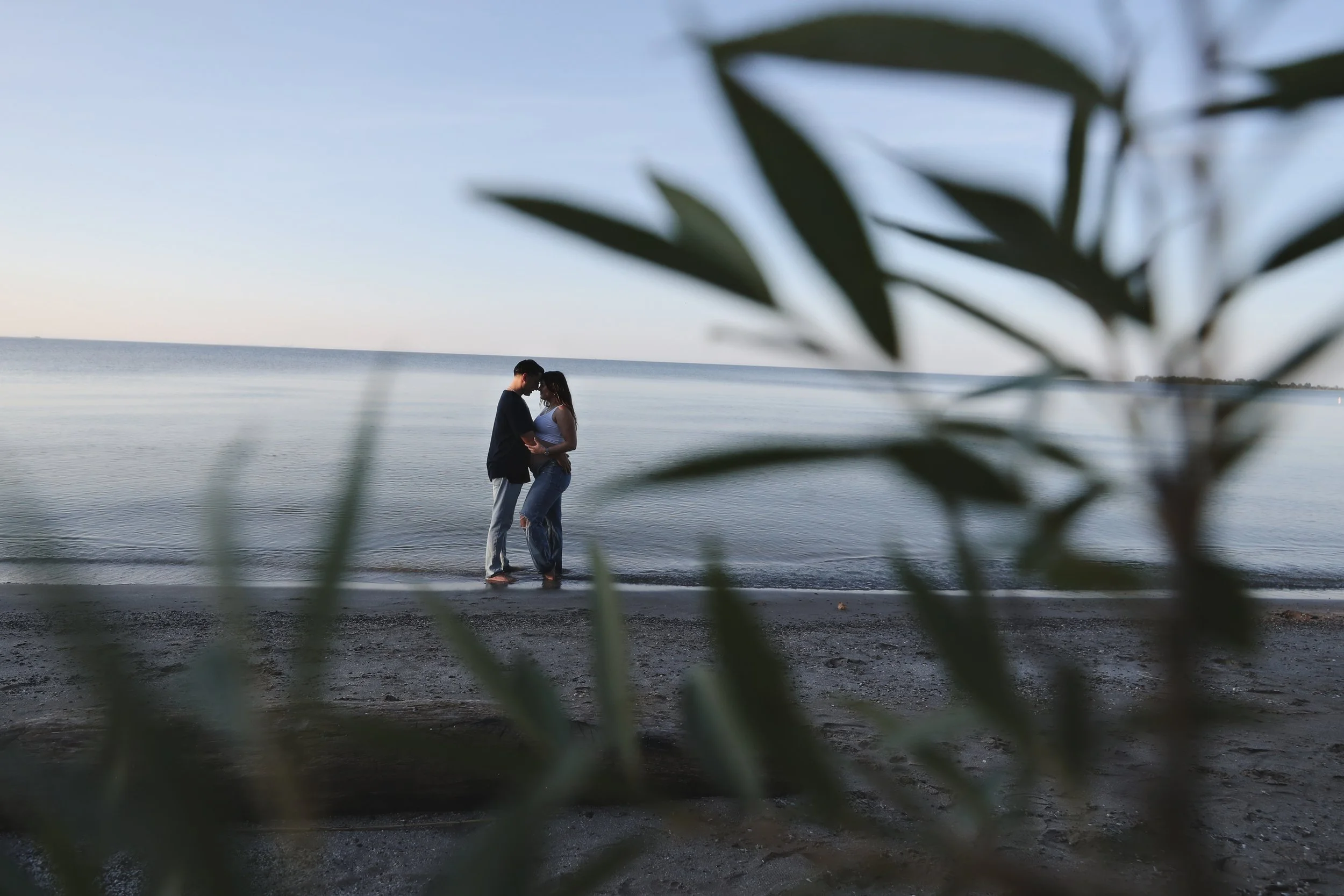 A couple stands close together on a beach, framed by blurred leaves in the foreground, with the water and sky in the background during sunset or early evening.